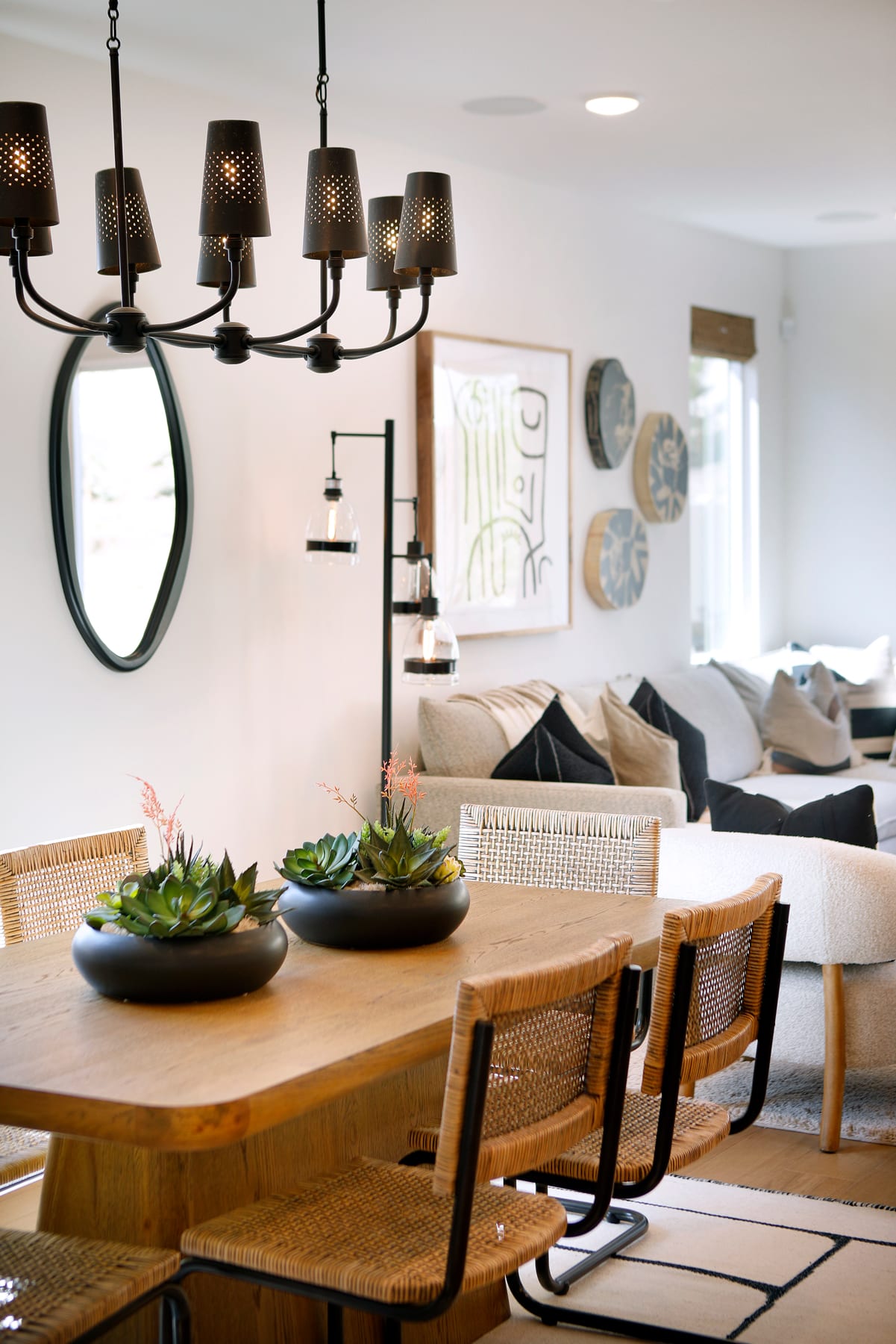A cozy and modern dining area with a wooden table, woven chairs, and a decorative chandelier, set against a backdrop of a comfortable living room with artwork on the walls.
