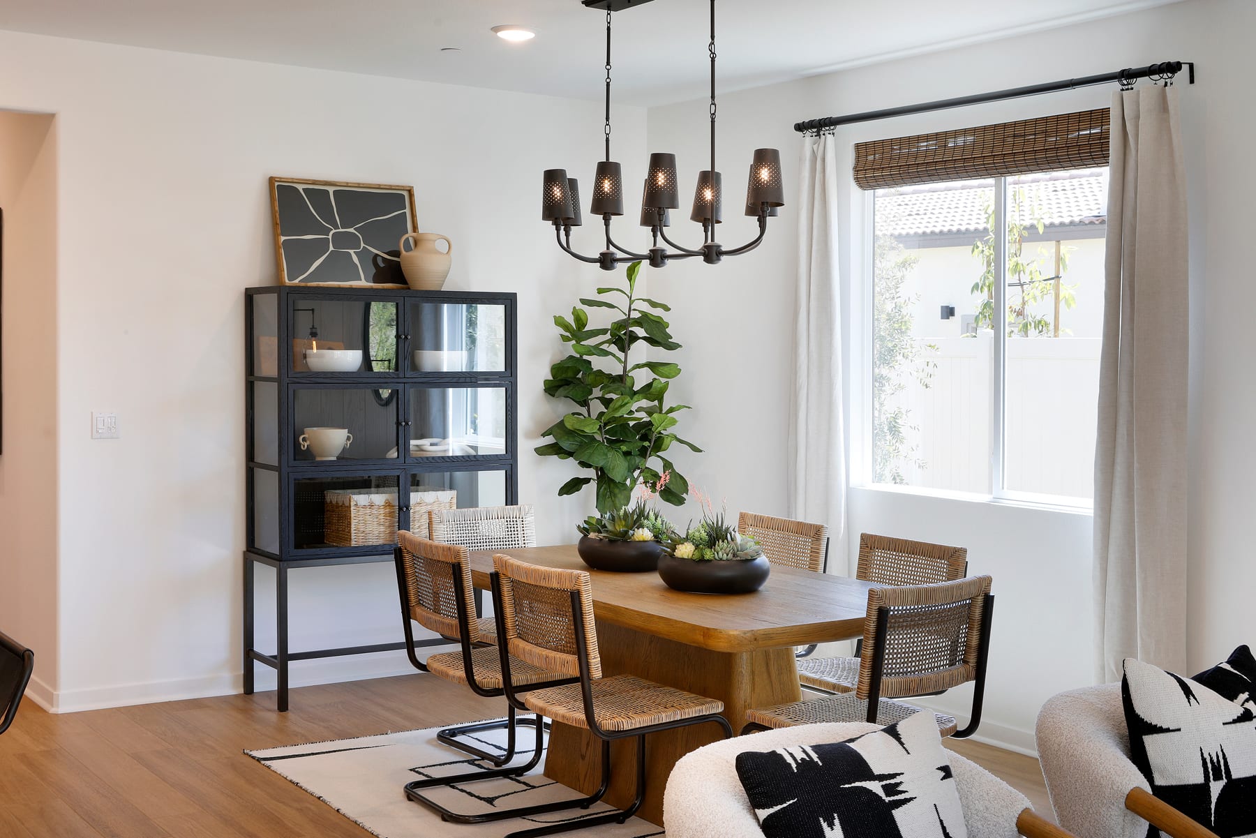 A cozy and well-decorated dining area with a wooden table, rattan chairs, a black metal shelving unit, and a large window providing natural light.