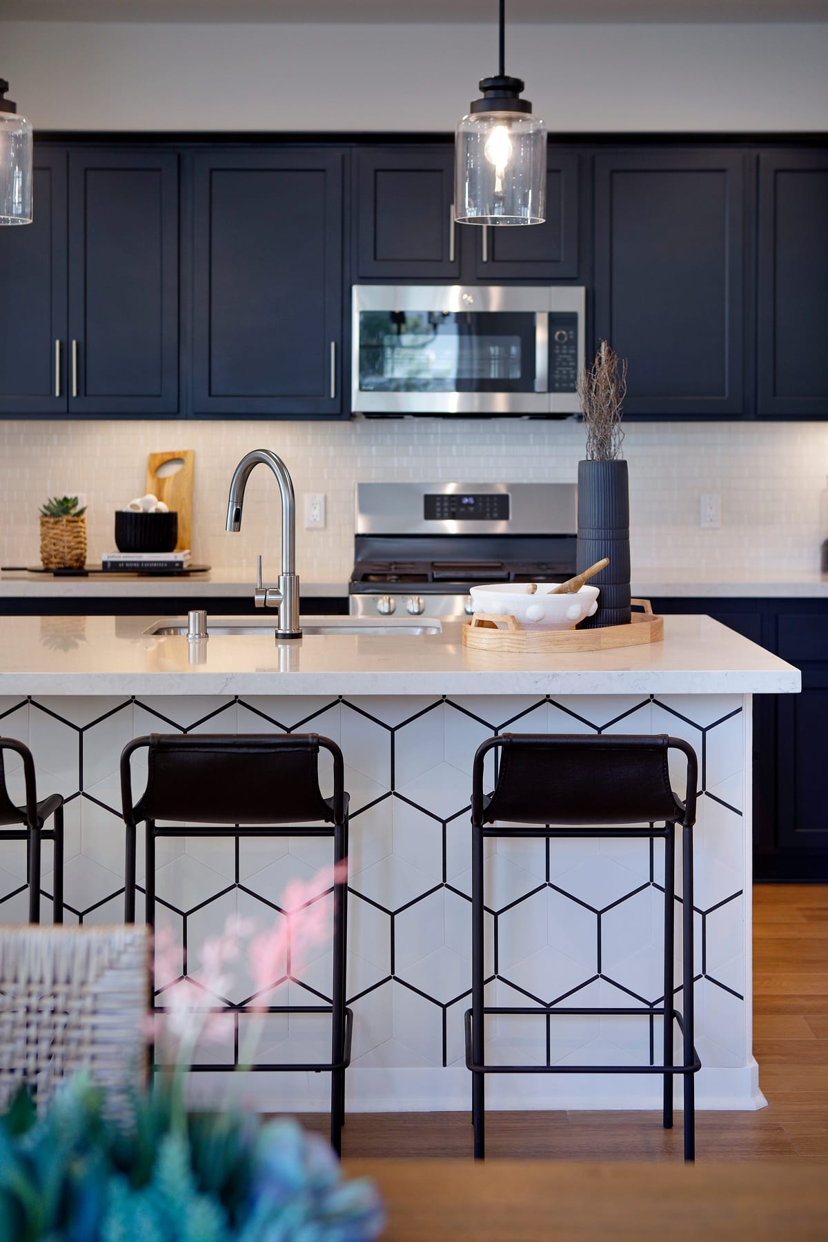A modern kitchen with dark cabinets, a geometric patterned island, and bar stools, featuring a microwave and other appliances on the countertop.