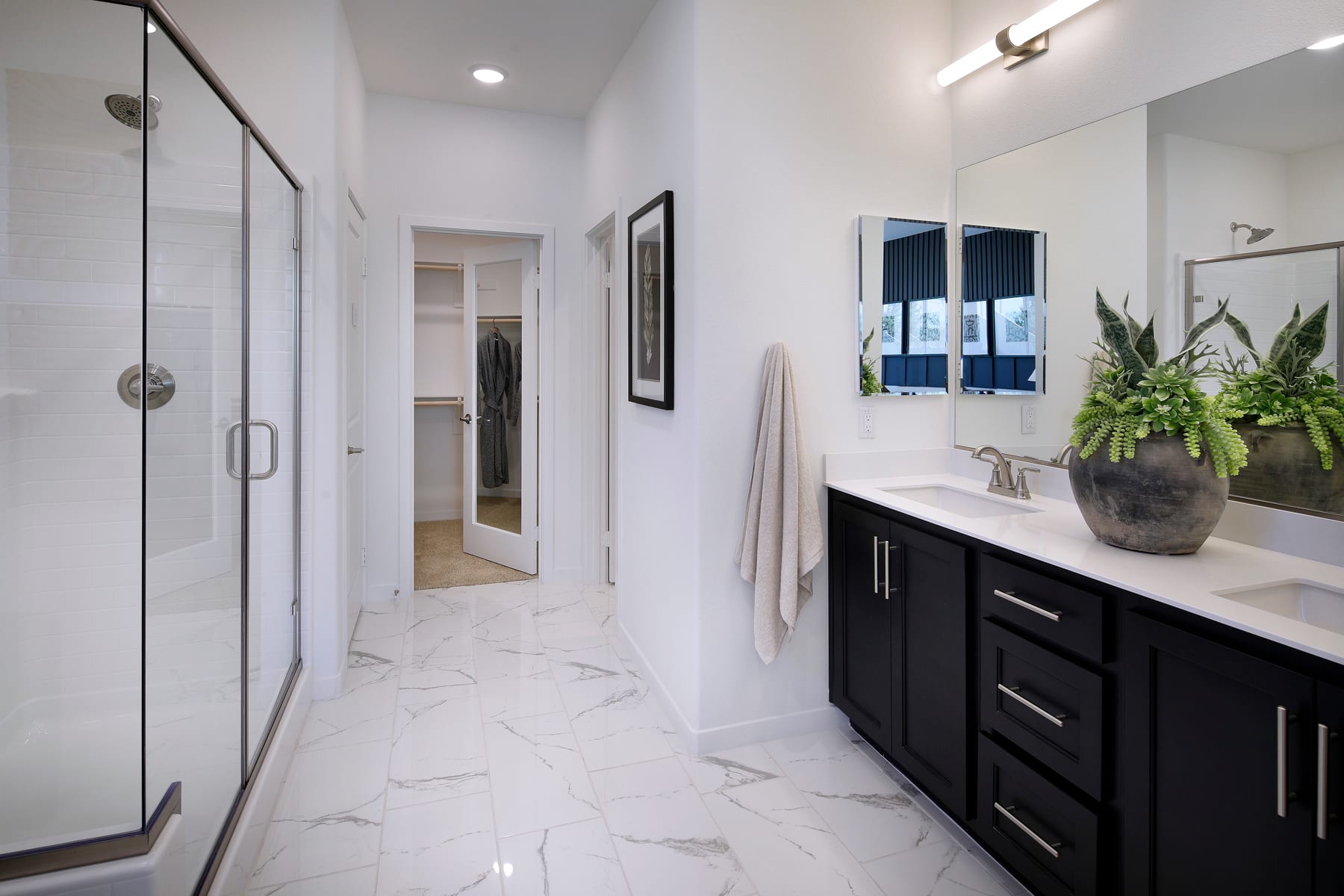 A modern and spacious bathroom with a marble-tiled floor, a dark wooden vanity, and a large mirror on the wall, complemented by potted plants and framed artwork.