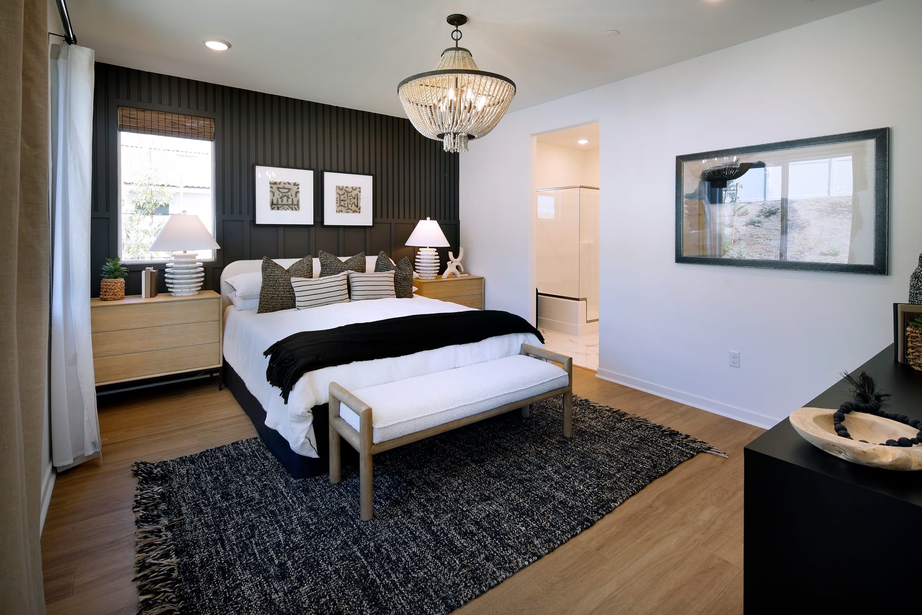 A cozy and modern bedroom with a large bed, wooden furniture, and a chandelier hanging from the ceiling, set against a dark accent wall with framed artwork.