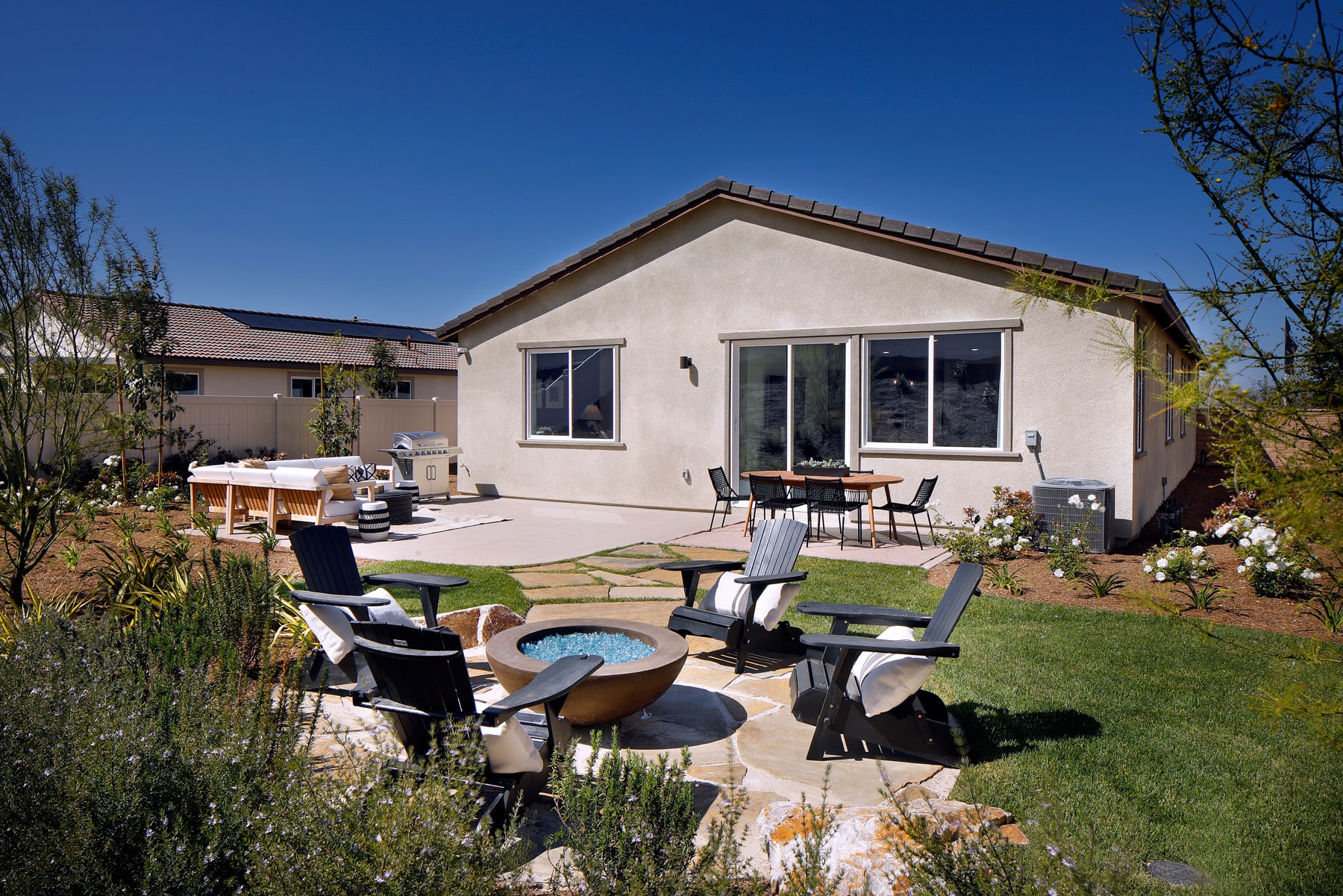 A cozy backyard with a patio set surrounded by lush greenery, against the backdrop of a well-maintained stucco house with large windows.