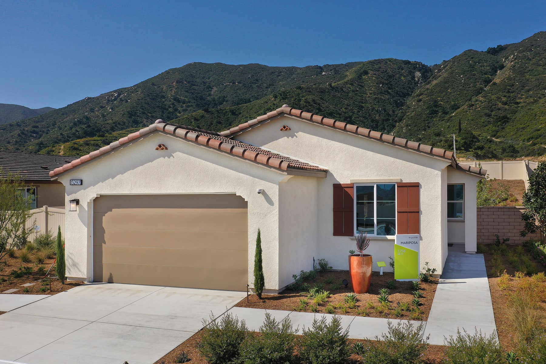 A single-story house with a beige exterior, a red tile roof, and a garage door sits in the foreground, surrounded by lush greenery and mountains in the background.