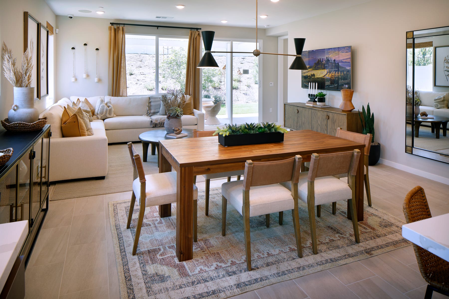A cozy and well-furnished dining area with a wooden table and chairs, surrounded by a living room with comfortable seating and decorative elements, all set against a backdrop of large windows providing natural light and a view of the outdoors.