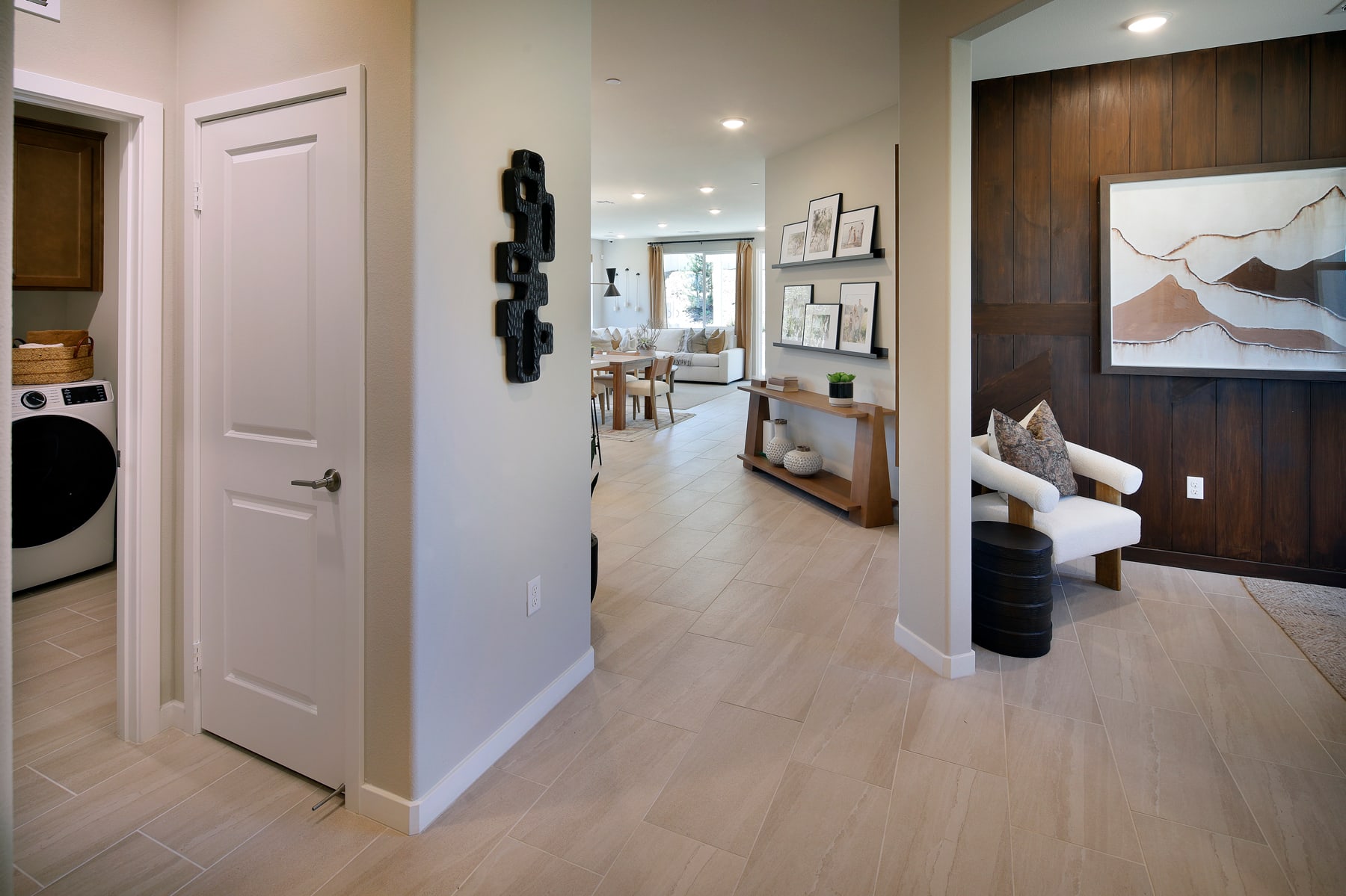 A bright and airy hallway with a white door, wooden floors, and artwork on the walls, leading to a cozy living room area with a comfortable-looking chair.