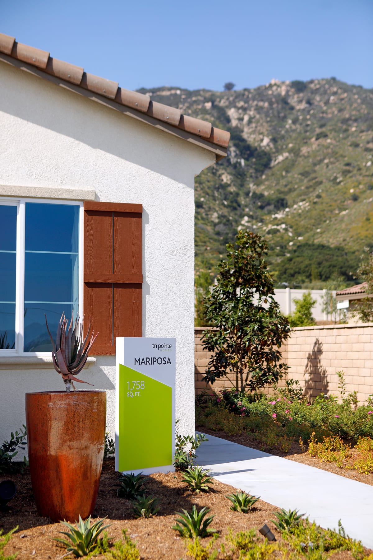 A modern white building with wooden shutters and a potted plant in the foreground, set against a backdrop of rolling hills and mountains.
