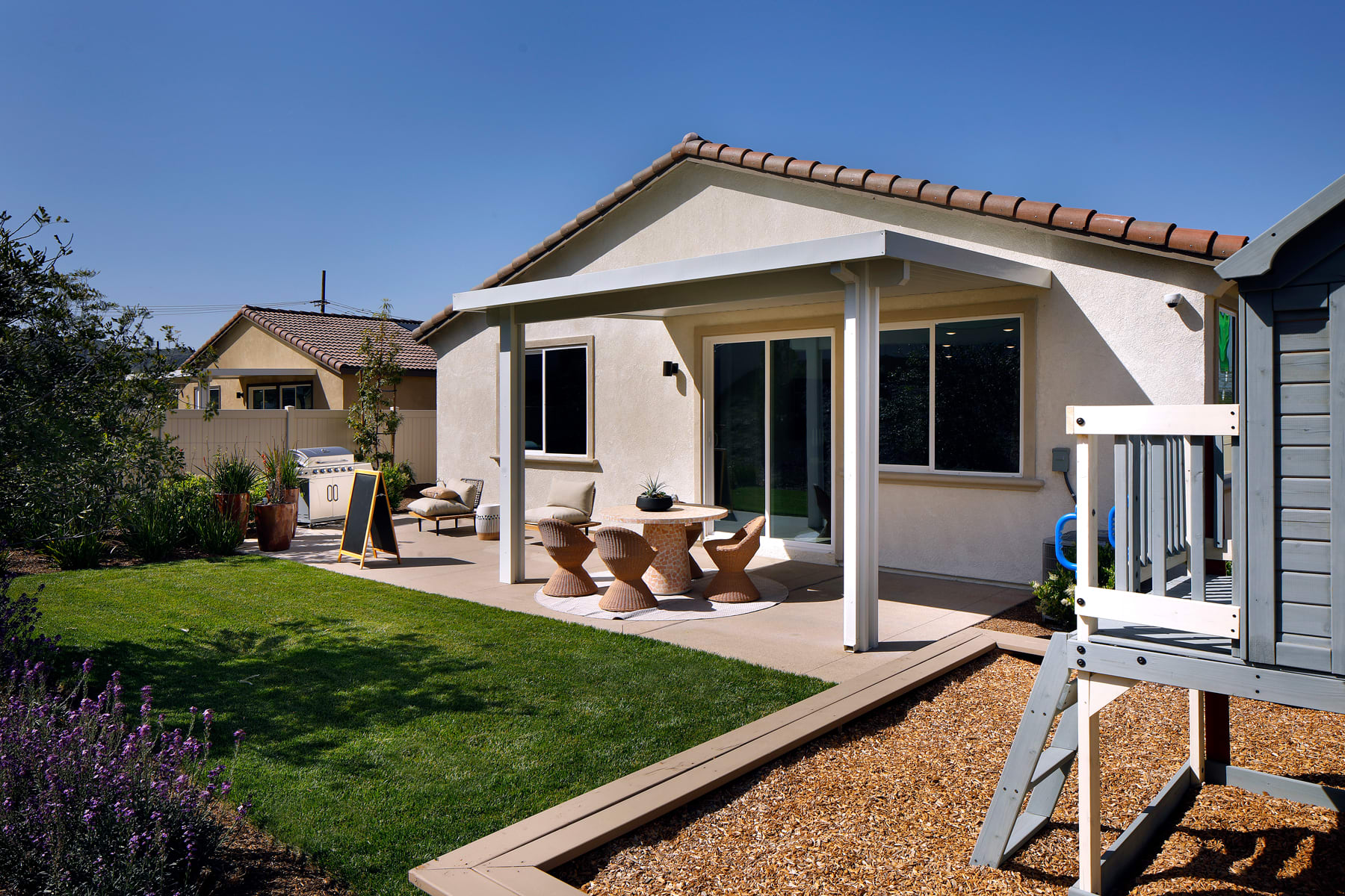 A cozy single-story house with a covered patio, surrounded by a well-maintained lawn and landscaping, including potted plants and a wooden deck in the foreground.