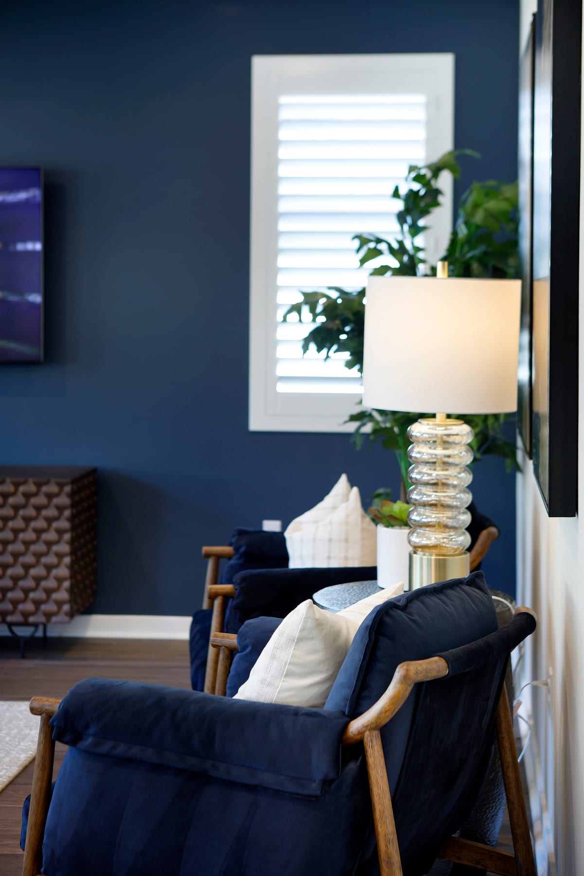 A cozy living room with a navy blue sofa, a wooden lamp, and potted plants against a dark blue wall with a framed window.