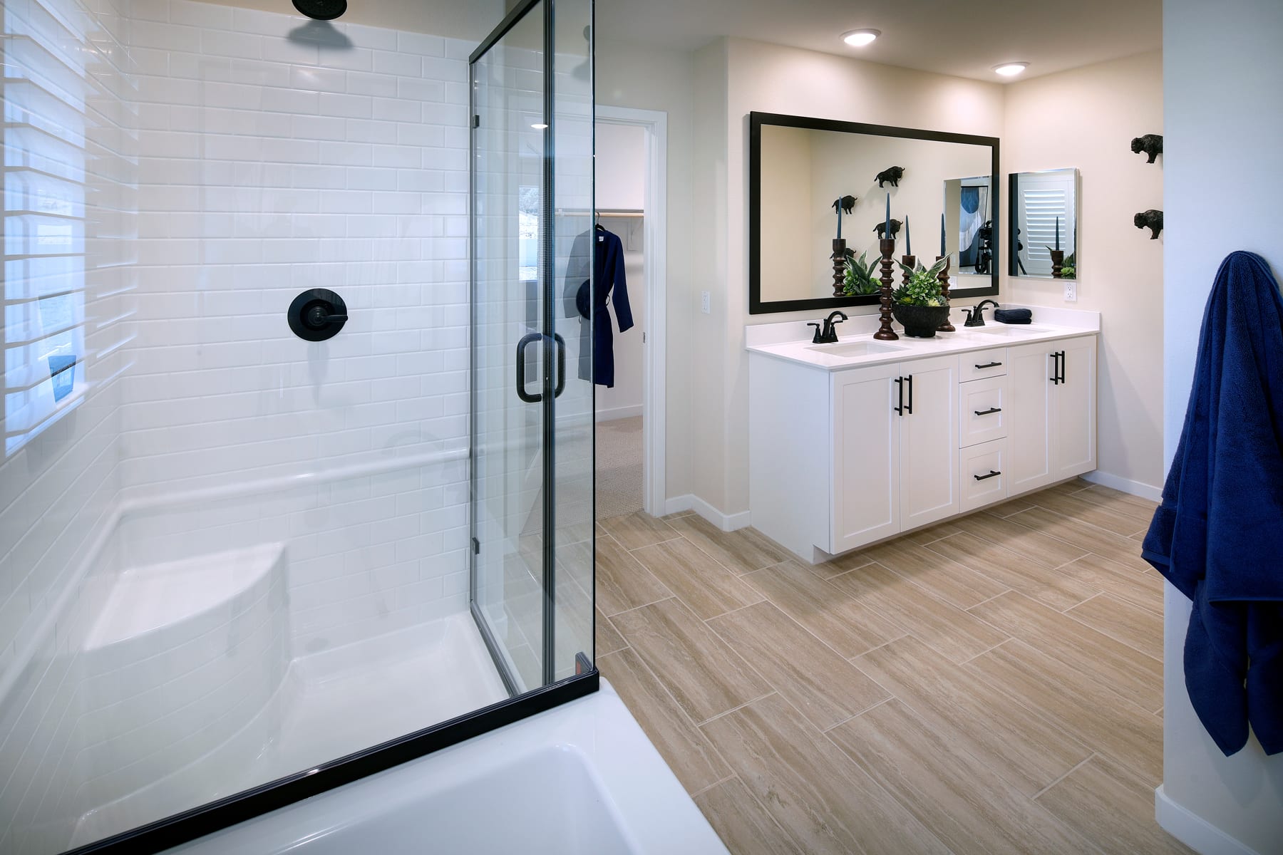 A modern and minimalist bathroom with a glass shower enclosure, a white vanity with drawers, and a large mirror on the wall. The floor is covered in light-colored wood-like tiles, creating a clean and airy atmosphere.