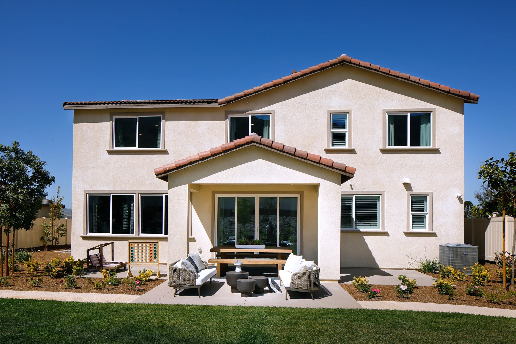 A two-story stucco house with a red tile roof, surrounded by a well-manicured lawn and landscaping, sits against a clear blue sky.
