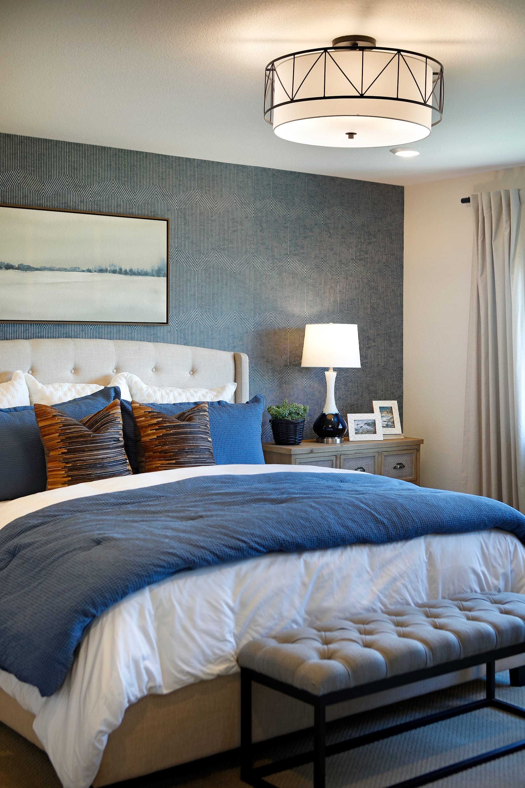 A cozy and modern bedroom with a large bed, patterned bedding, and a stylish overhead light fixture, set against a backdrop of a textured gray wall and a framed landscape artwork.