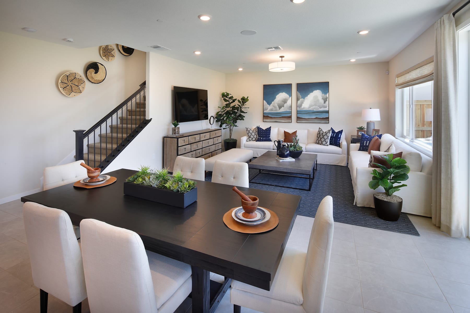 A modern and stylish living room with a dining area, featuring a dark wooden table, white chairs, and various decorative elements such as plants and artwork on the walls.