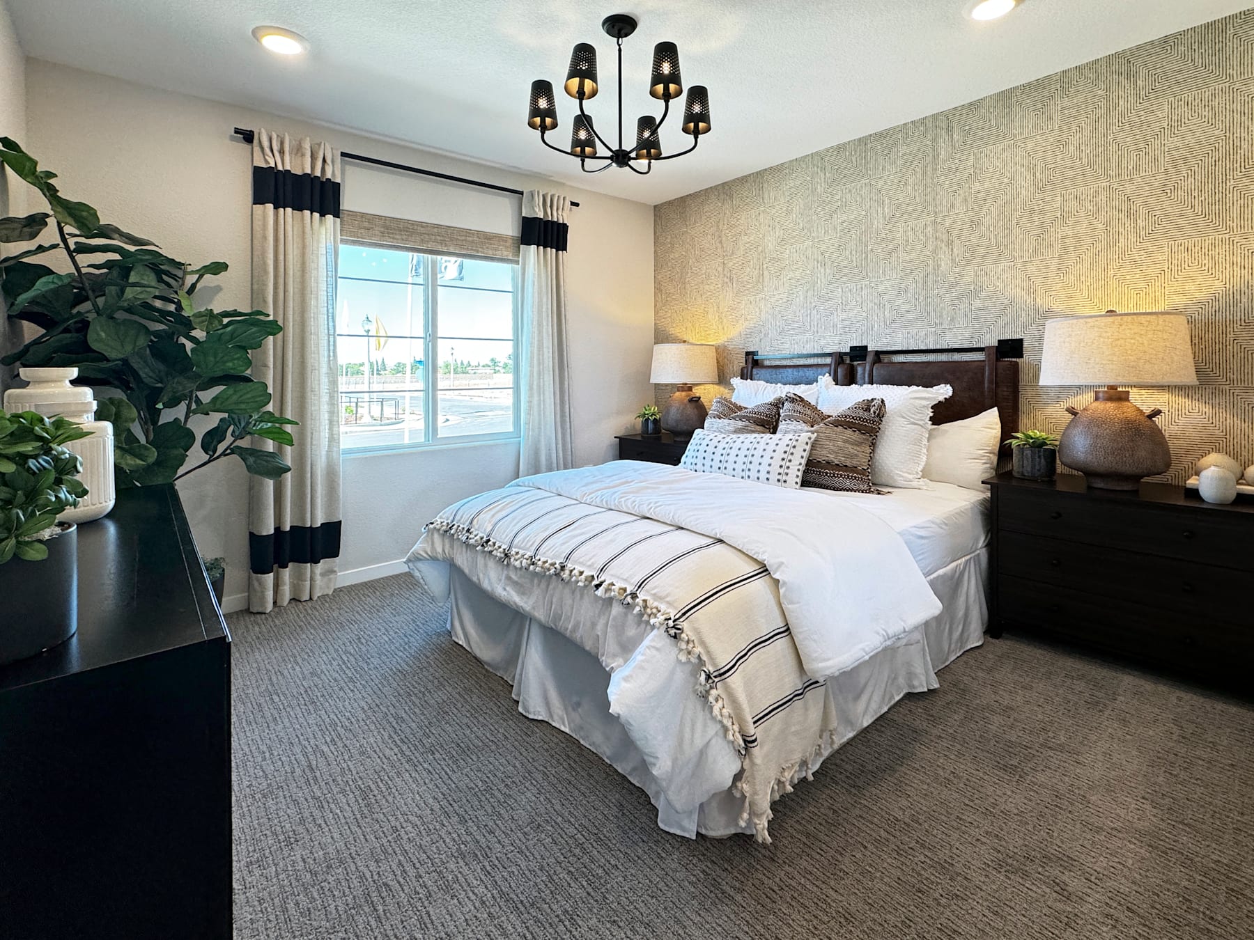 A cozy and well-appointed bedroom with a large bed, a chandelier, and a potted plant in the foreground, set against a backdrop of curtained windows and a neutral-toned decor.