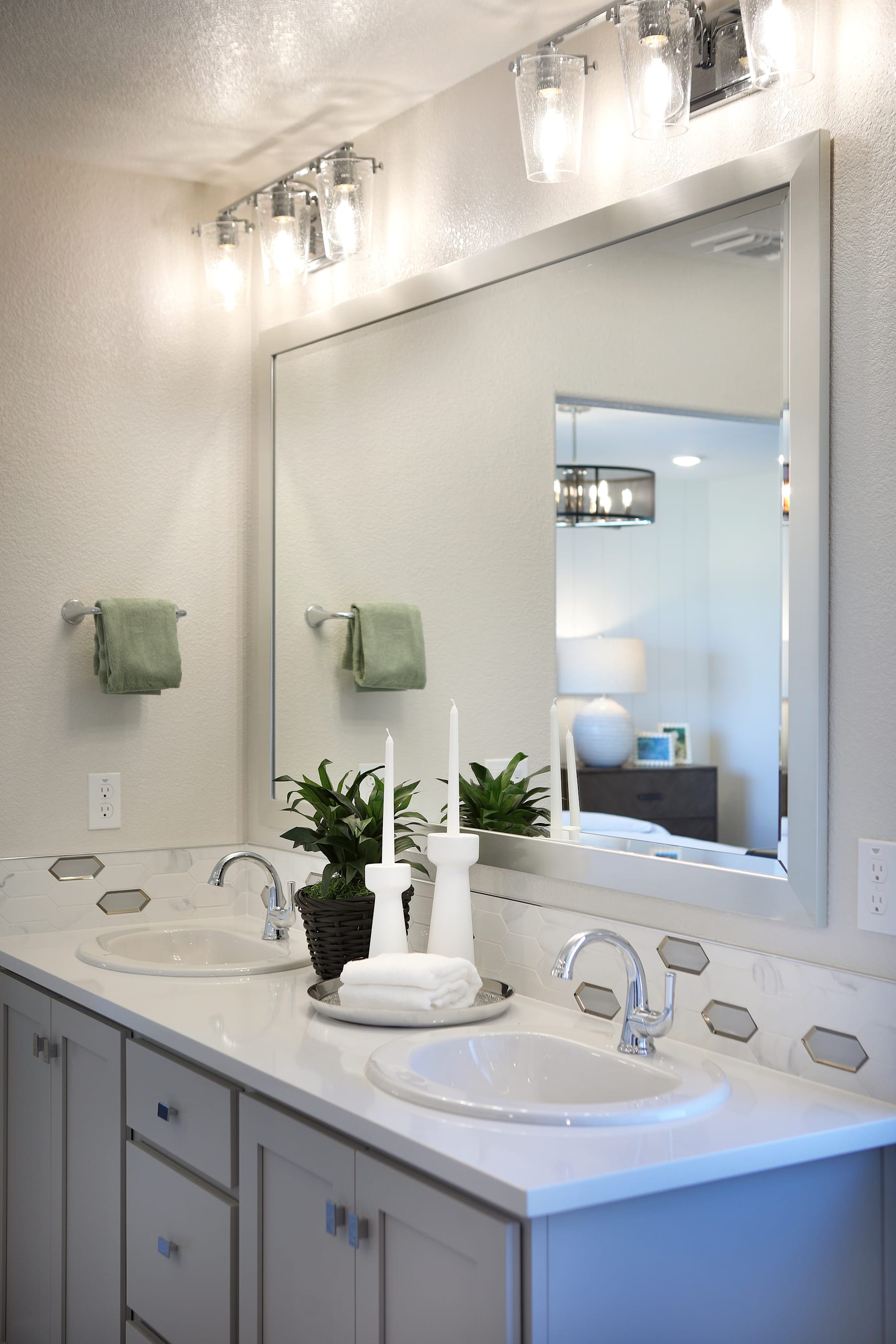 A modern and well-lit bathroom with a double vanity, white countertops, and a large mirror on the wall. The room features green accents and potted plants, creating a clean and refreshing atmosphere.