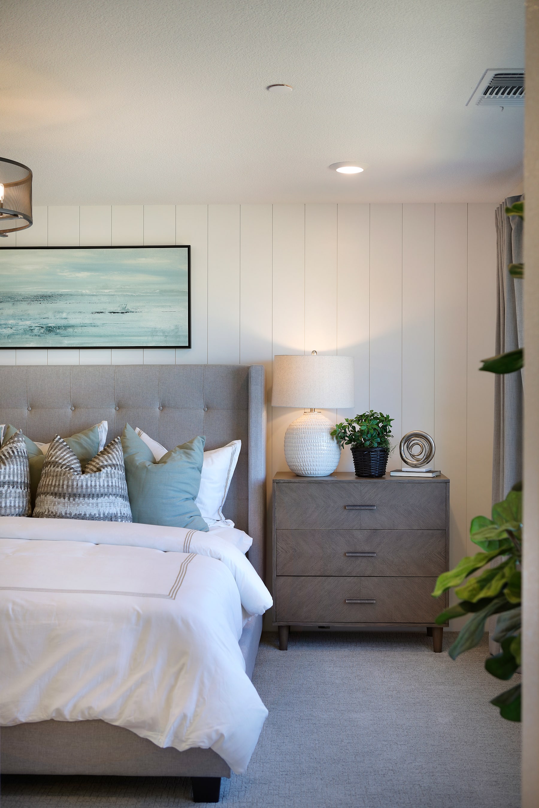 A cozy and inviting bedroom with a large bed, gray upholstered headboard, and a wooden dresser, complemented by a framed artwork depicting a serene ocean scene and potted plants adding a touch of greenery.