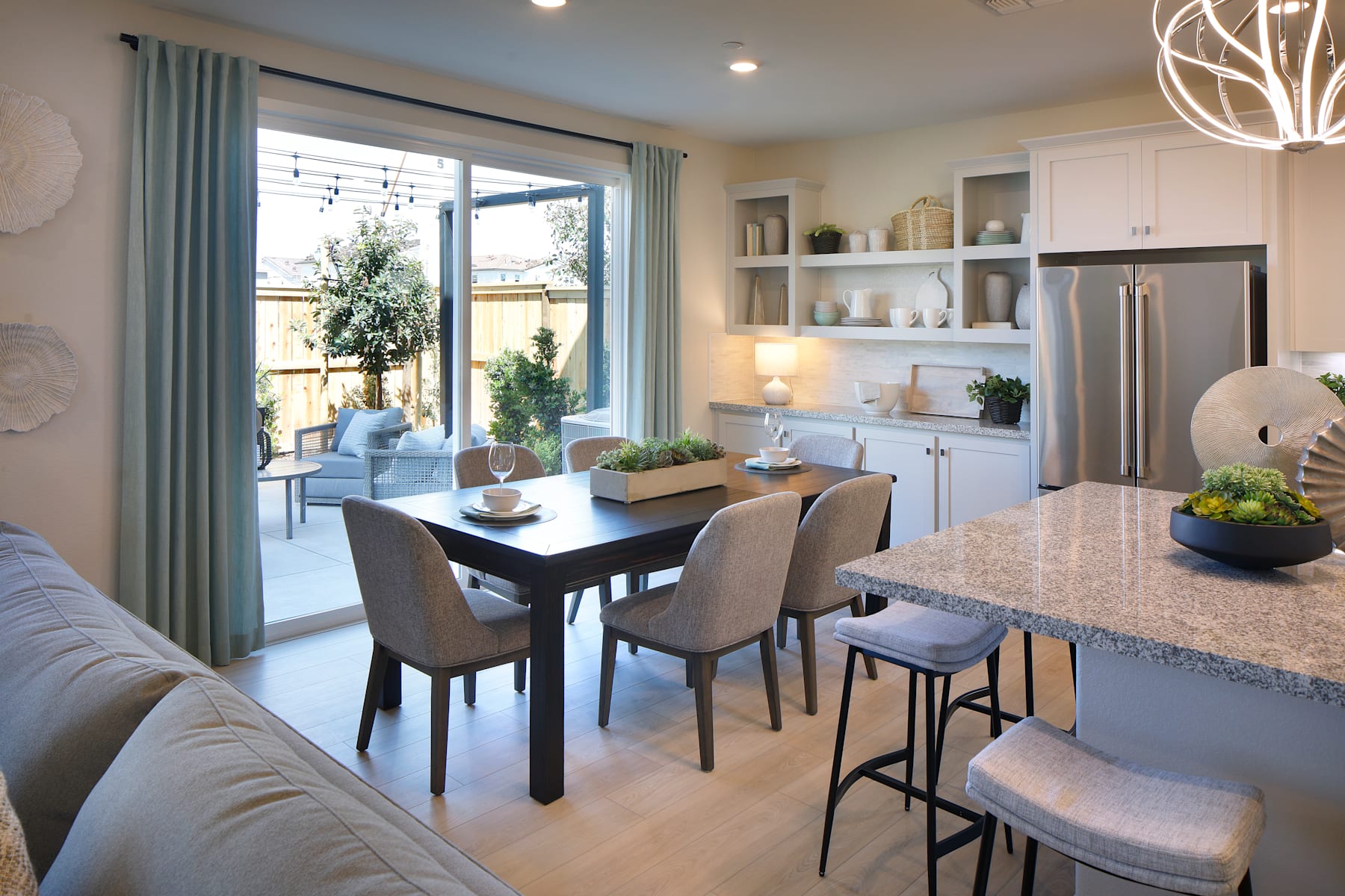 A modern and cozy kitchen-dining area with a large window overlooking a patio, featuring a wooden dining table, upholstered chairs, and built-in shelving displaying decorative items.