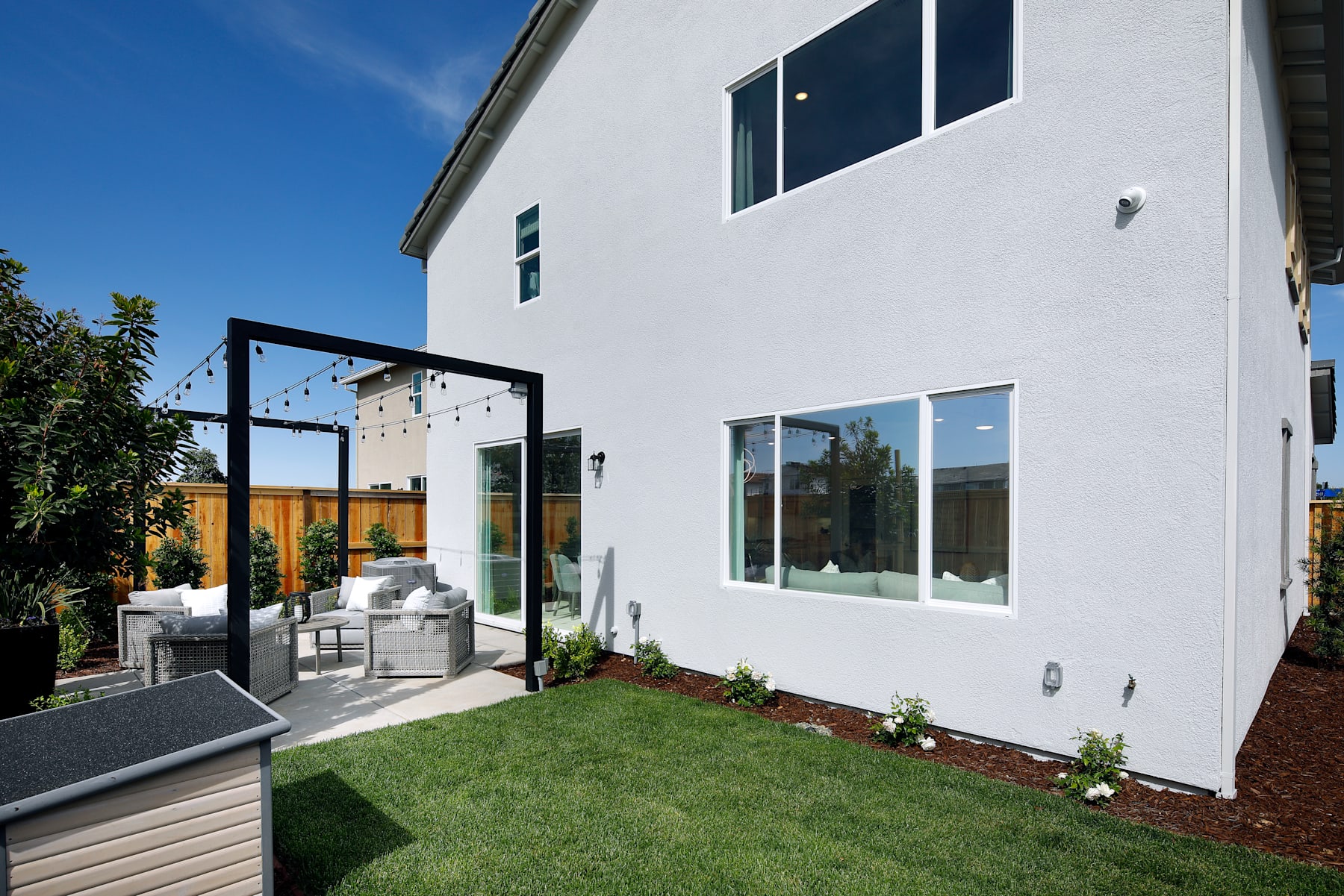 A modern, white two-story house with large windows and a covered patio area in the foreground, surrounded by a well-maintained lawn and landscaping.