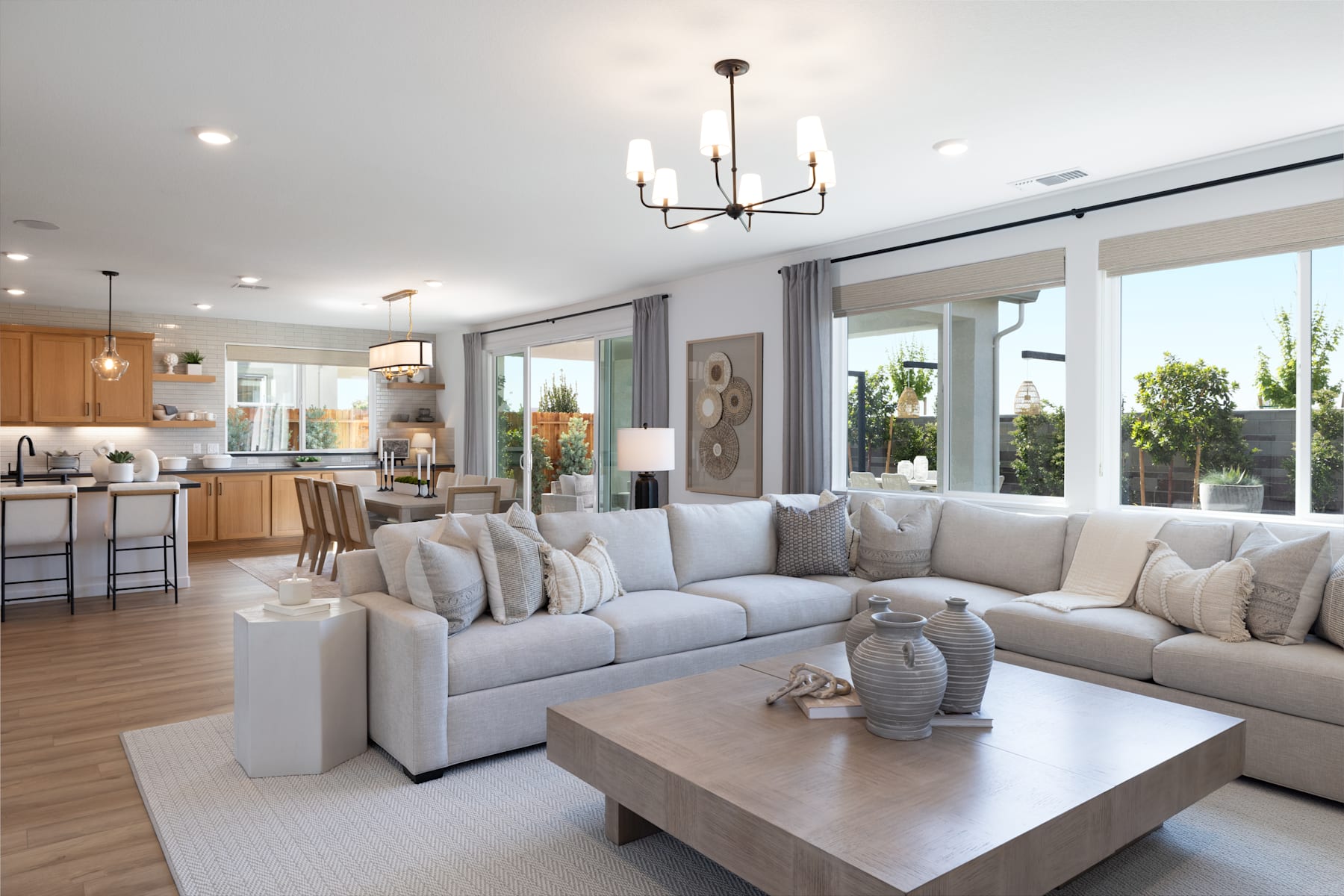 A spacious and modern living room with a large sectional sofa, a wooden coffee table, and a chandelier hanging from the ceiling. The room has large windows that provide natural light and a view of the outdoor scenery.