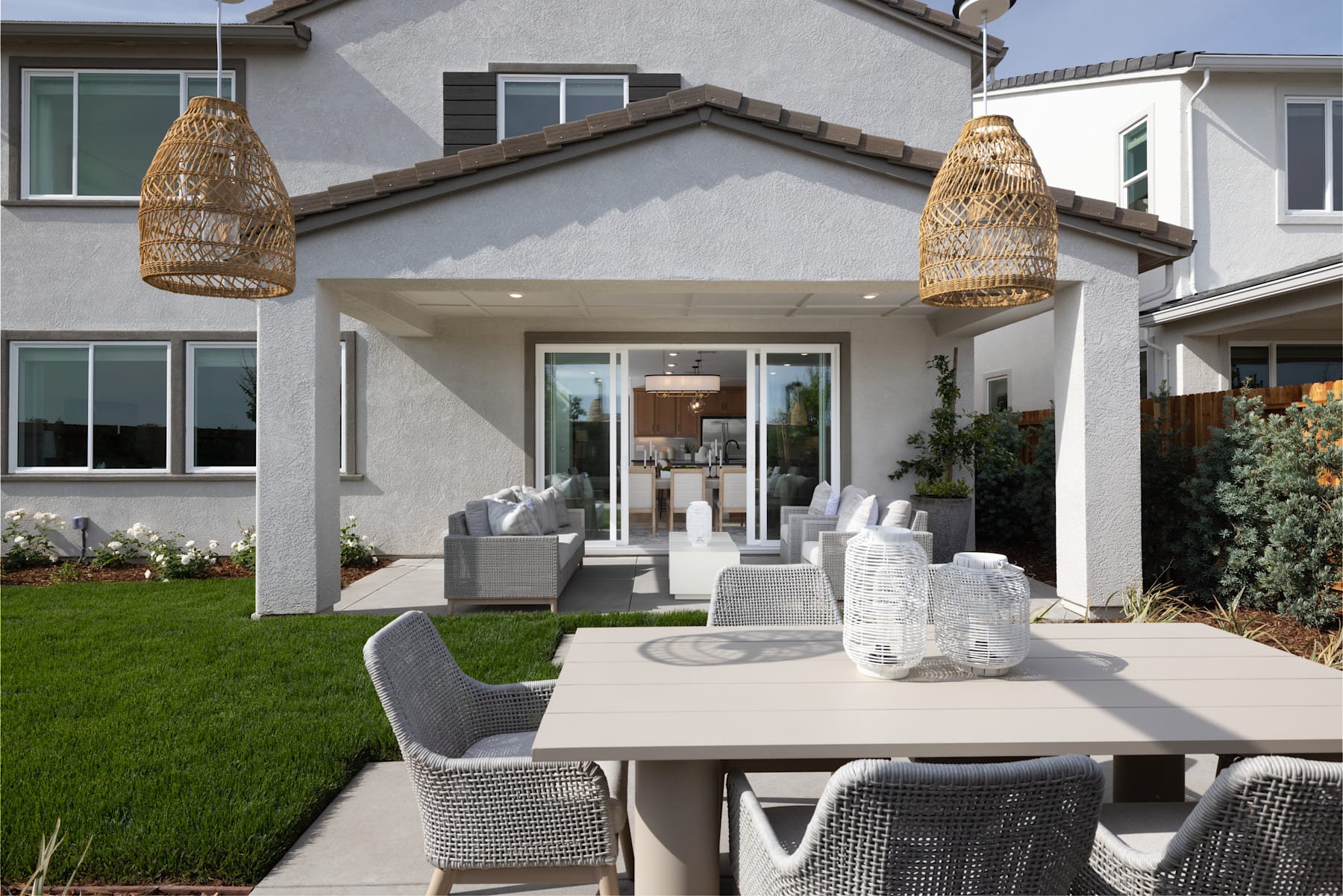 A modern, well-designed outdoor living space with a dining table, comfortable seating, and hanging woven light fixtures, set against a backdrop of a residential building with a manicured lawn.