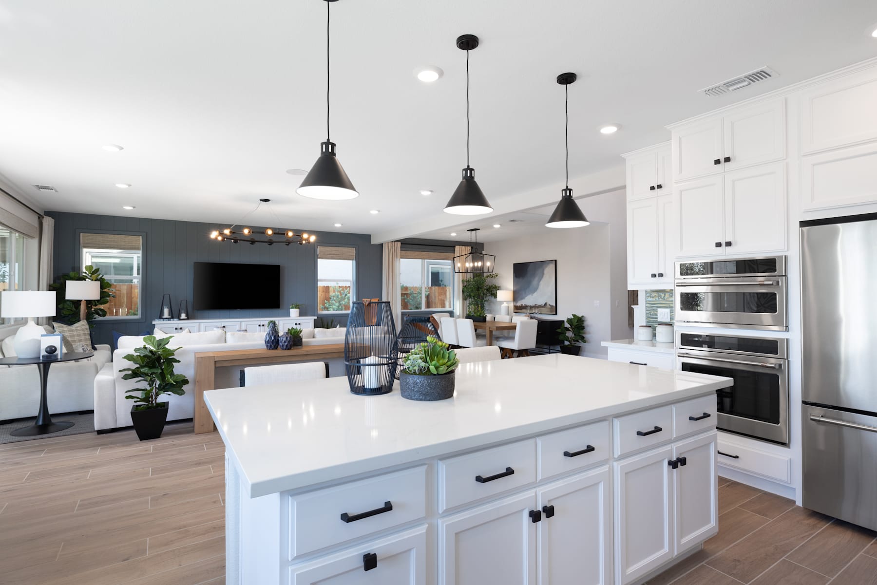 A modern, open-concept kitchen with a large white island, pendant lighting, and a mix of white and wood-toned cabinetry, set against a backdrop of a living room area with a television.