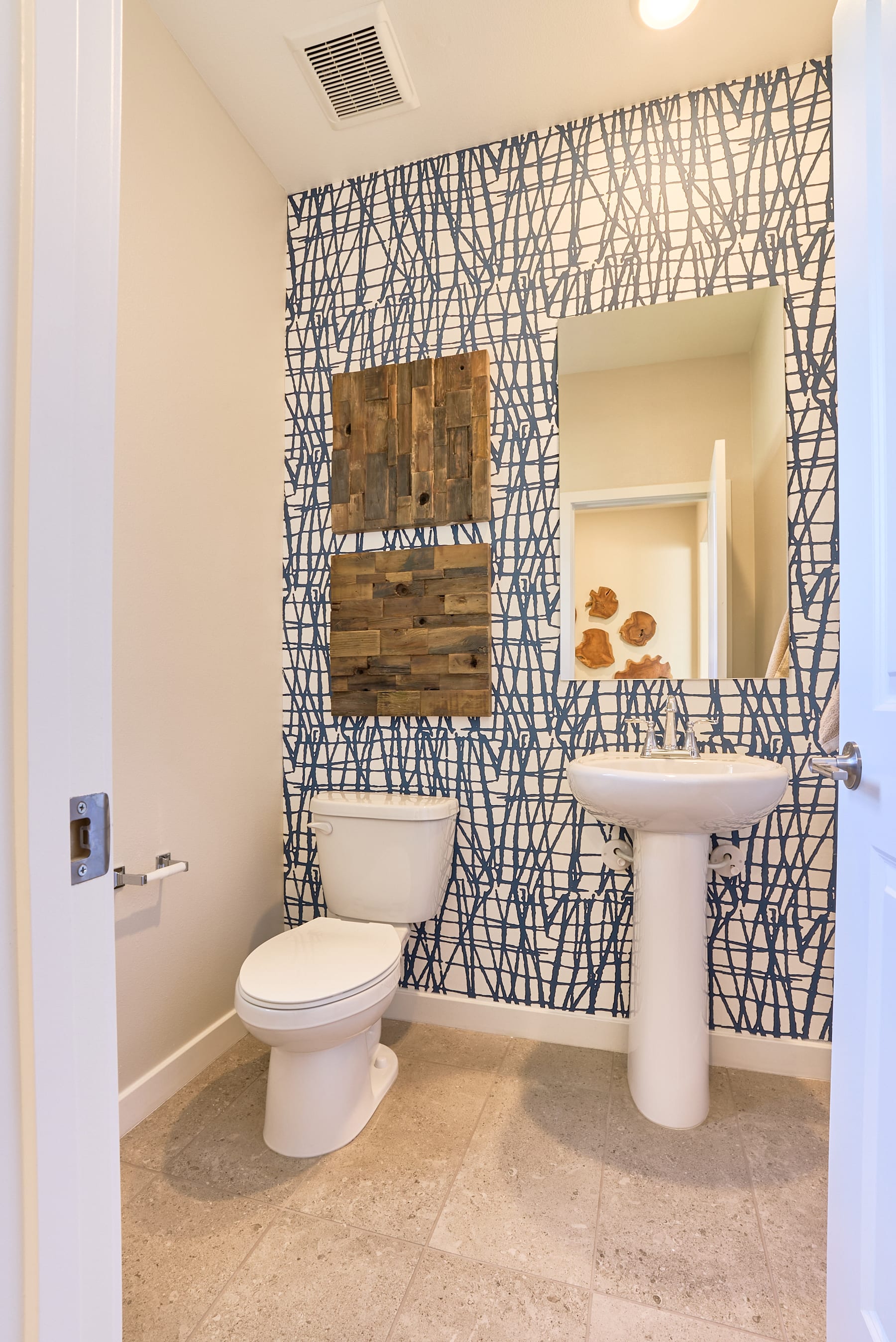 A modern bathroom with a patterned wallpaper, a wooden wall-mounted shelf, and a pedestal sink.
