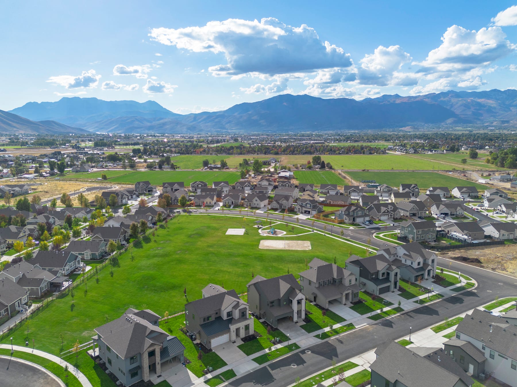 A sprawling residential neighborhood nestled among lush green fields, with towering mountains visible in the distance under a picturesque cloudy sky.
