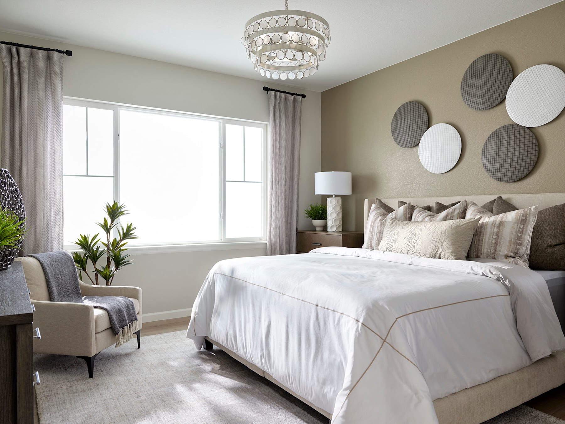 A cozy and well-designed bedroom with a large bed, plush bedding, and a decorative chandelier overhead. The room features neutral tones, textured wall decor, and a comfortable seating area near the window.