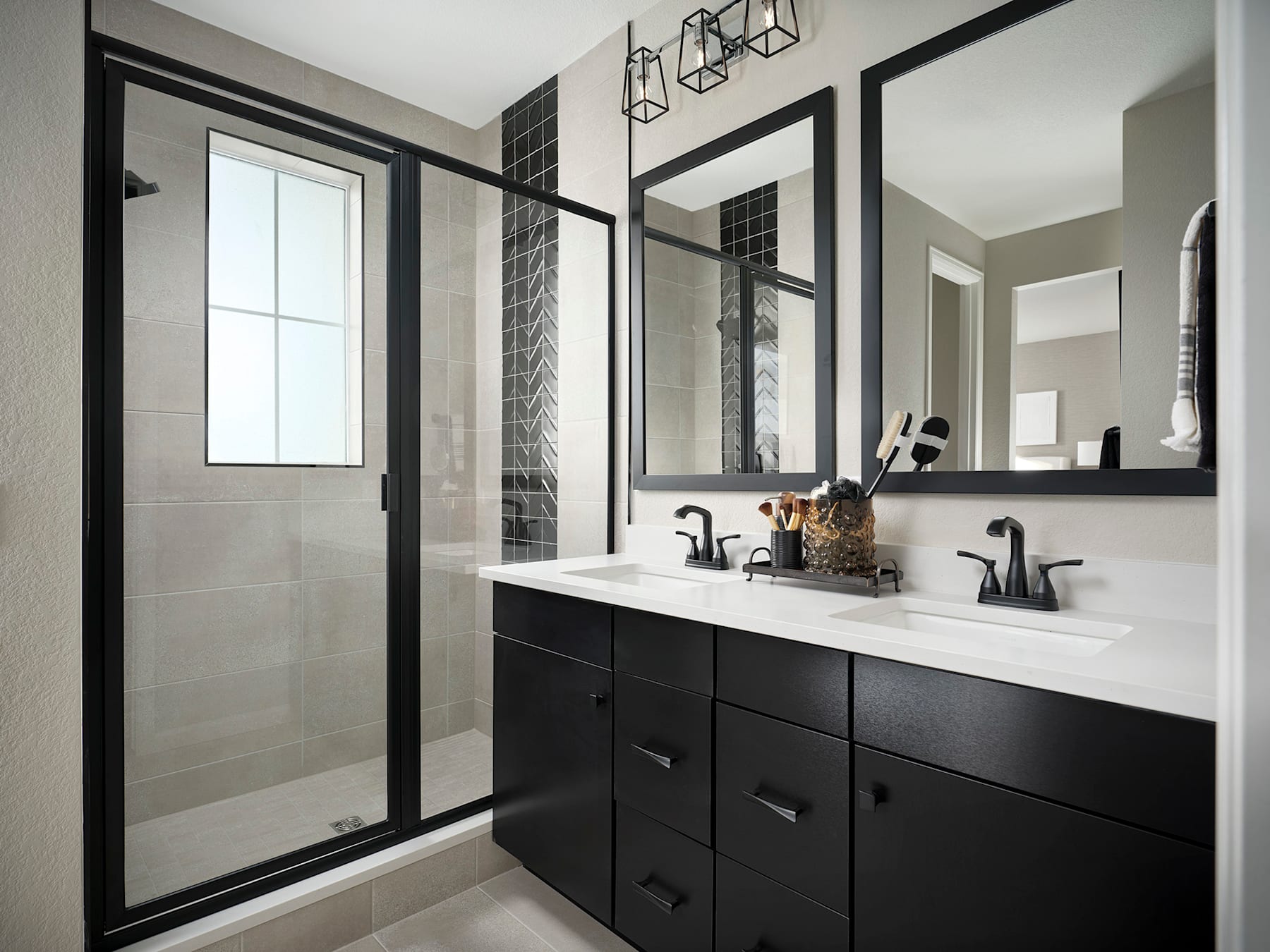 A modern bathroom with a black vanity, white countertop, and a large mirror framed in black. The room features a glass shower enclosure and a window, creating a bright and airy atmosphere.