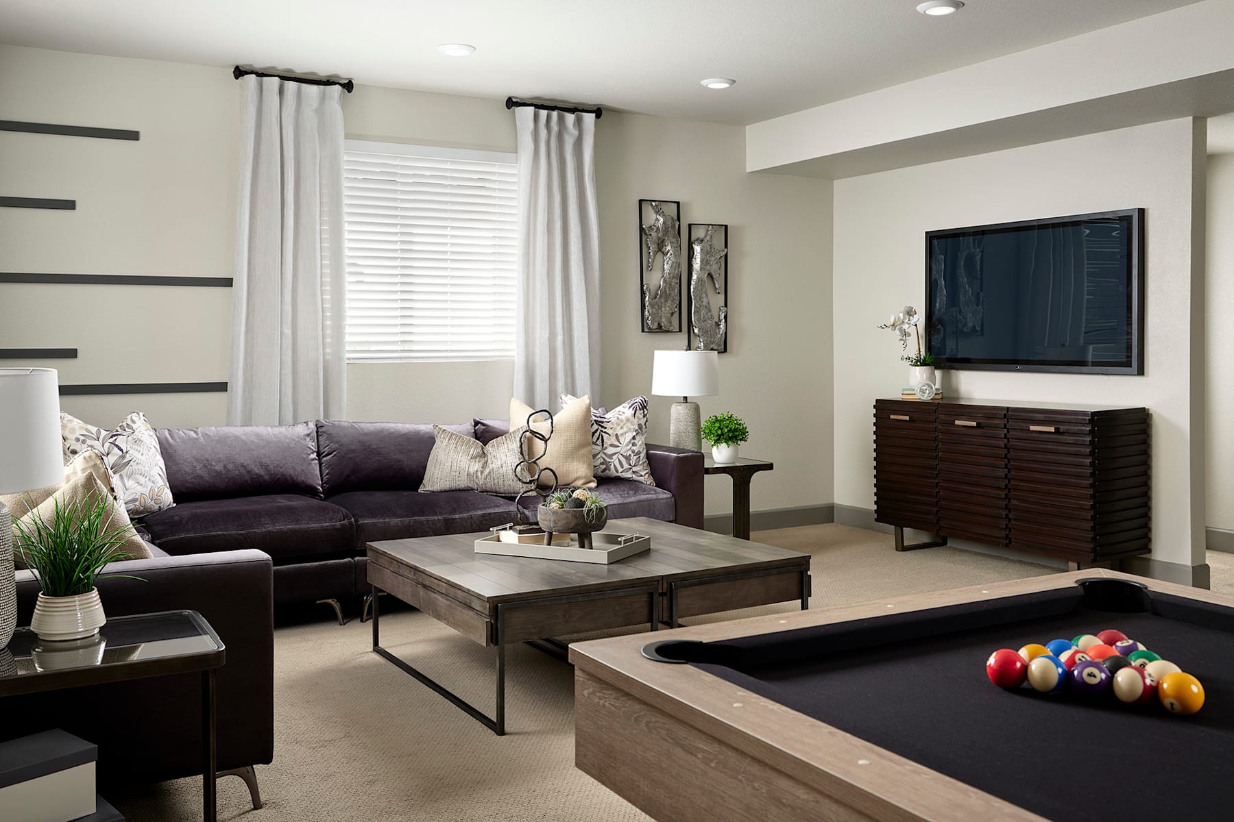A modern and stylish living room with a dark sofa, a wooden coffee table, and a television stand with decorative elements, set against a neutral-toned wall with curtains and artwork.