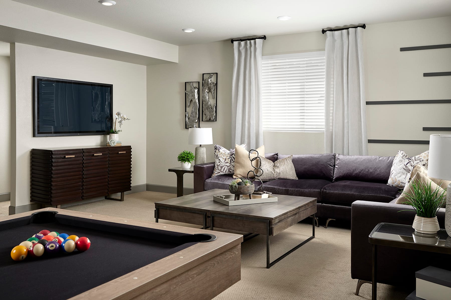 A modern and stylish living room with a large television, dark furniture, and decorative accents, including a coffee table with various objects and a potted plant.