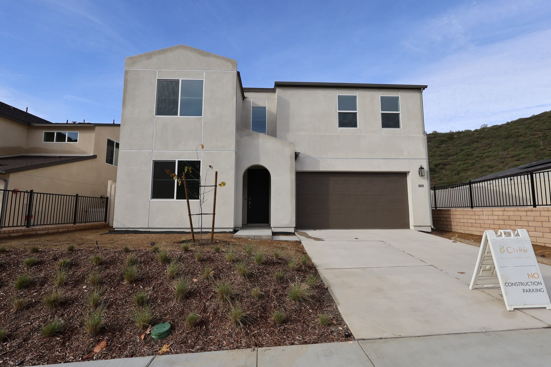 A modern two-story house with a garage, surrounded by a grassy area and other residential buildings in the background, set against a clear blue sky.
