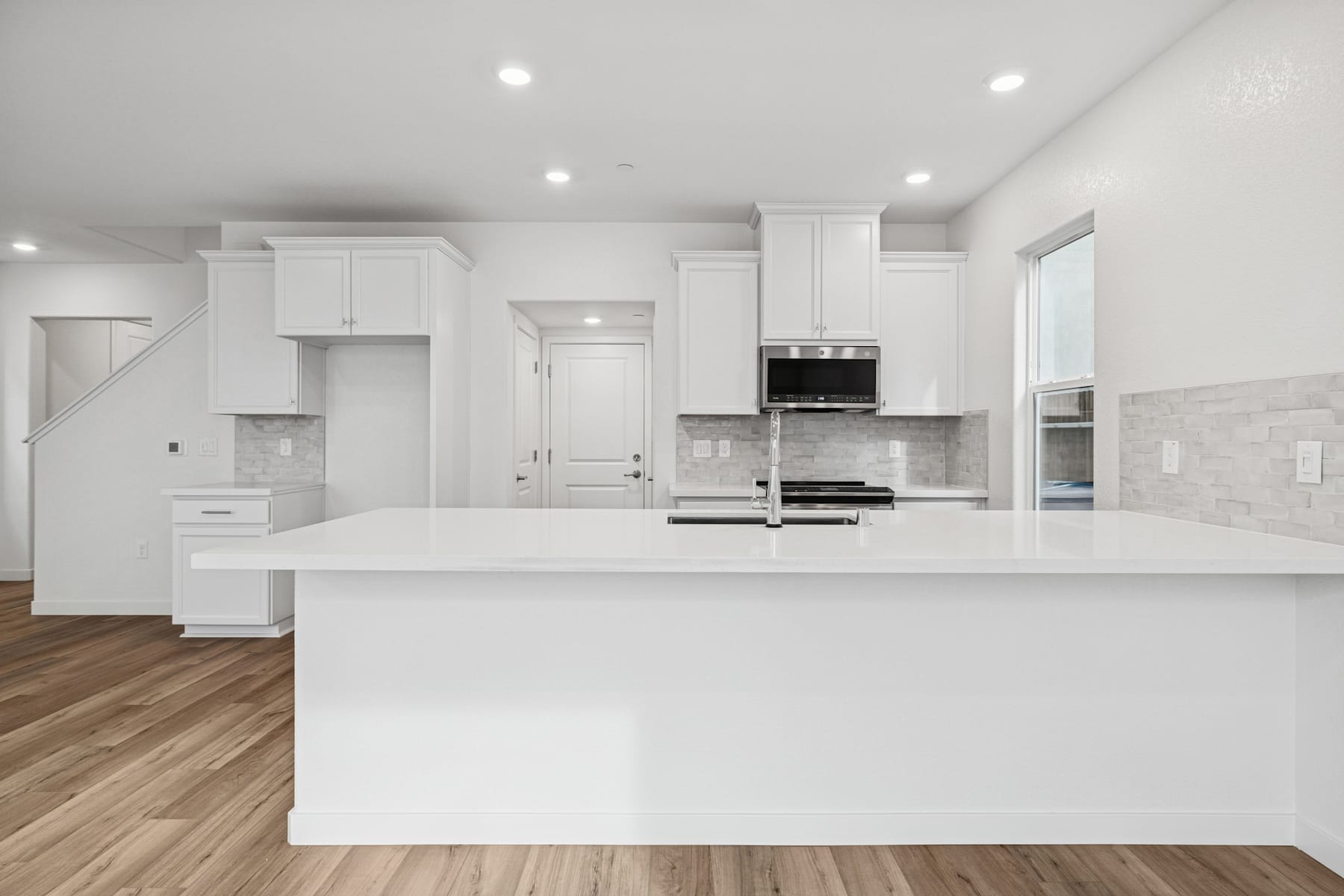 A modern, all-white kitchen with a large central island, wooden flooring, and recessed lighting fixtures.