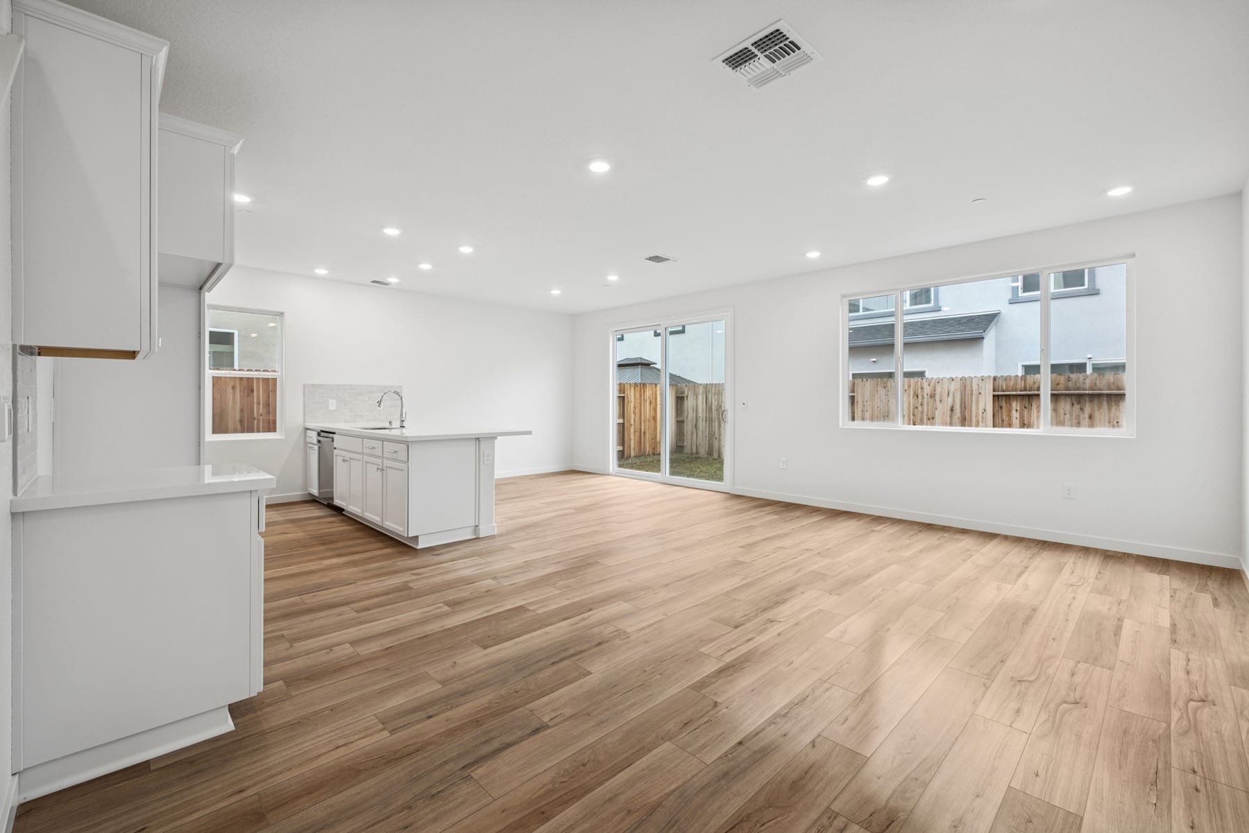 A spacious, well-lit room with a hardwood floor, white walls, and a kitchen area visible in the background.