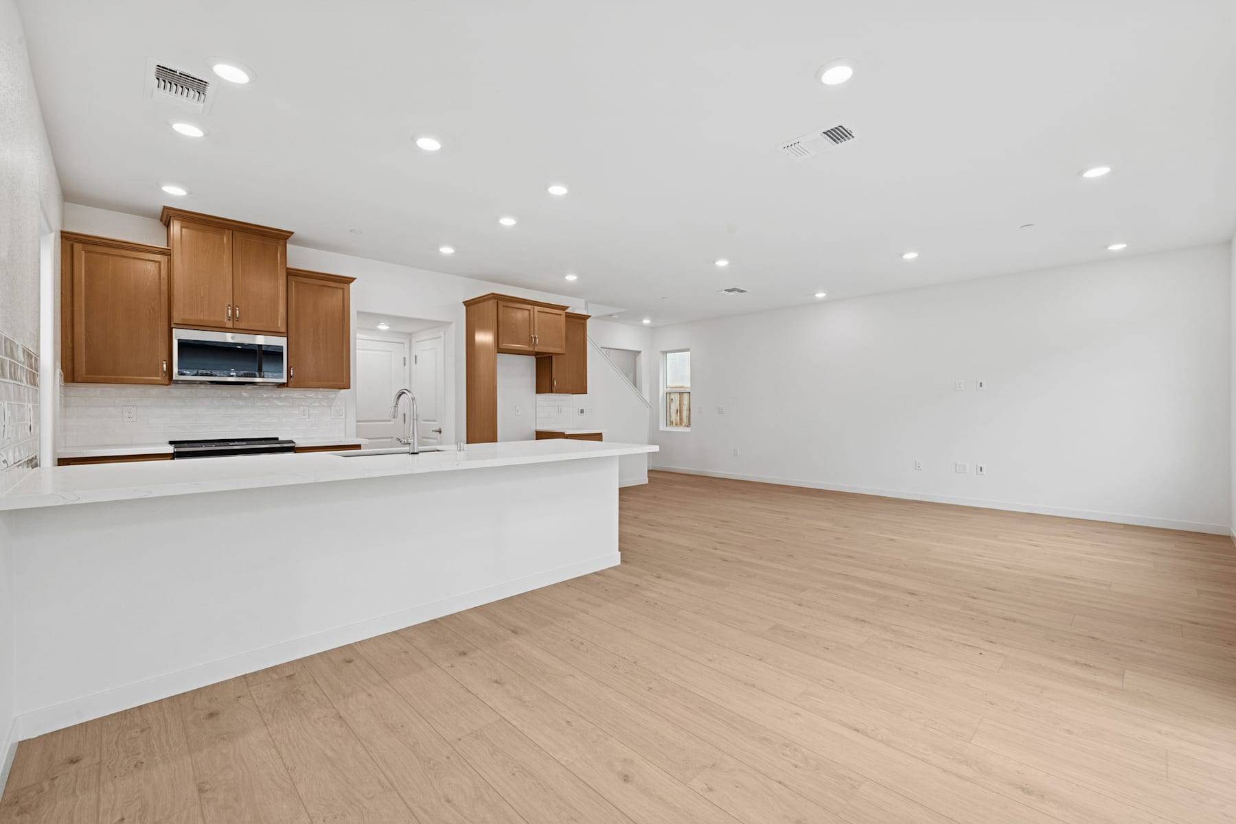 A modern, open-concept kitchen and living space with white walls, wooden cabinets, and a hardwood floor.