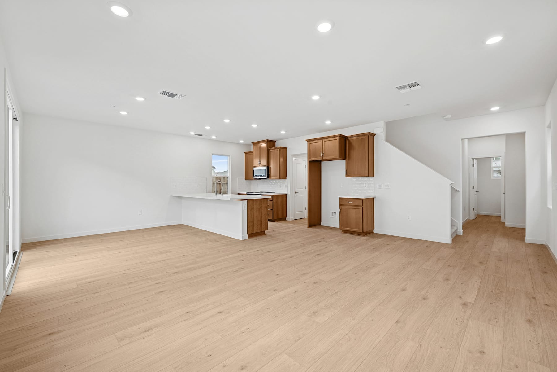 A spacious, open-concept living area with light-colored hardwood floors, white walls, and wooden kitchen cabinets.