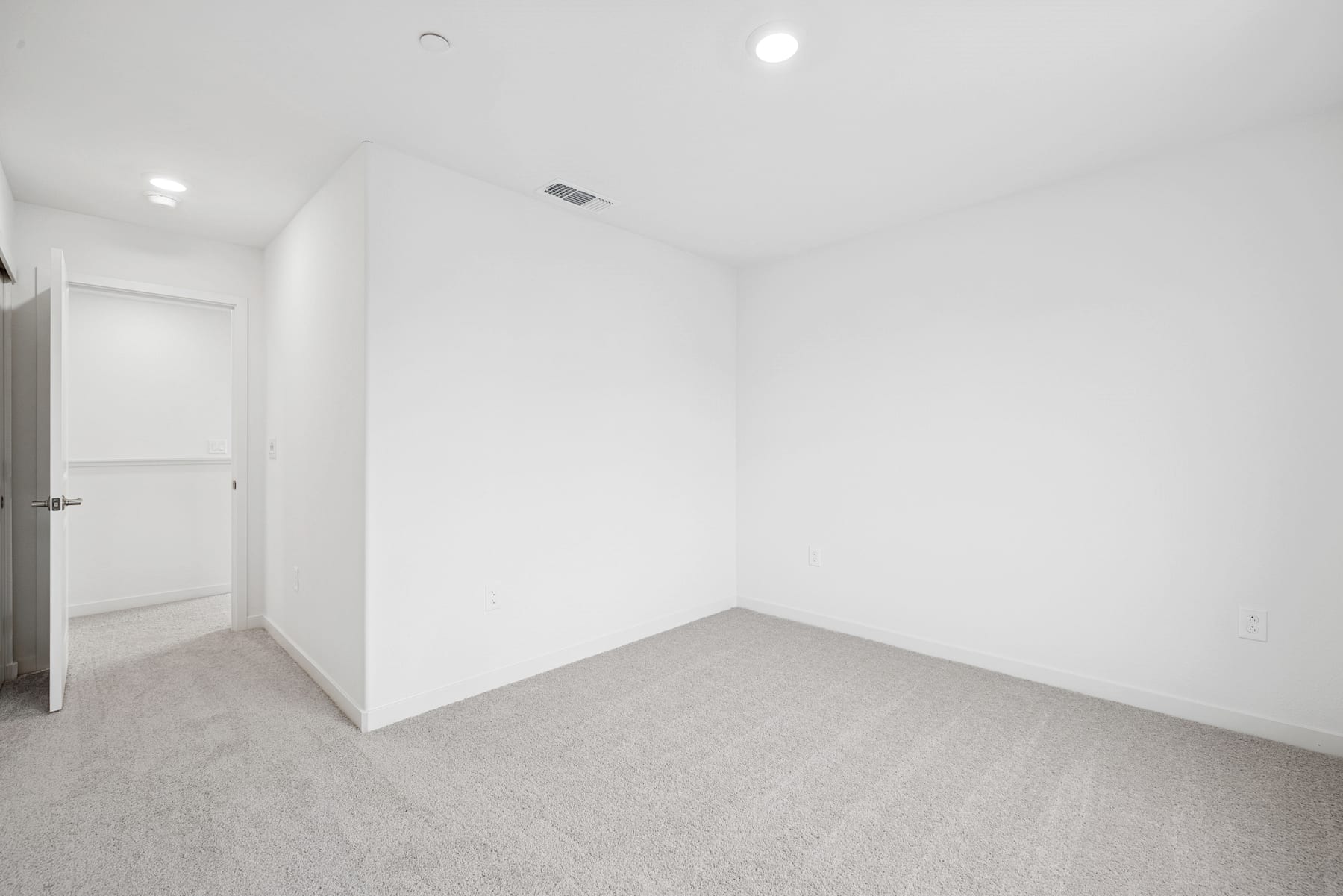 A minimalist, white-walled room with a gray carpeted floor and a closed door visible in the background.