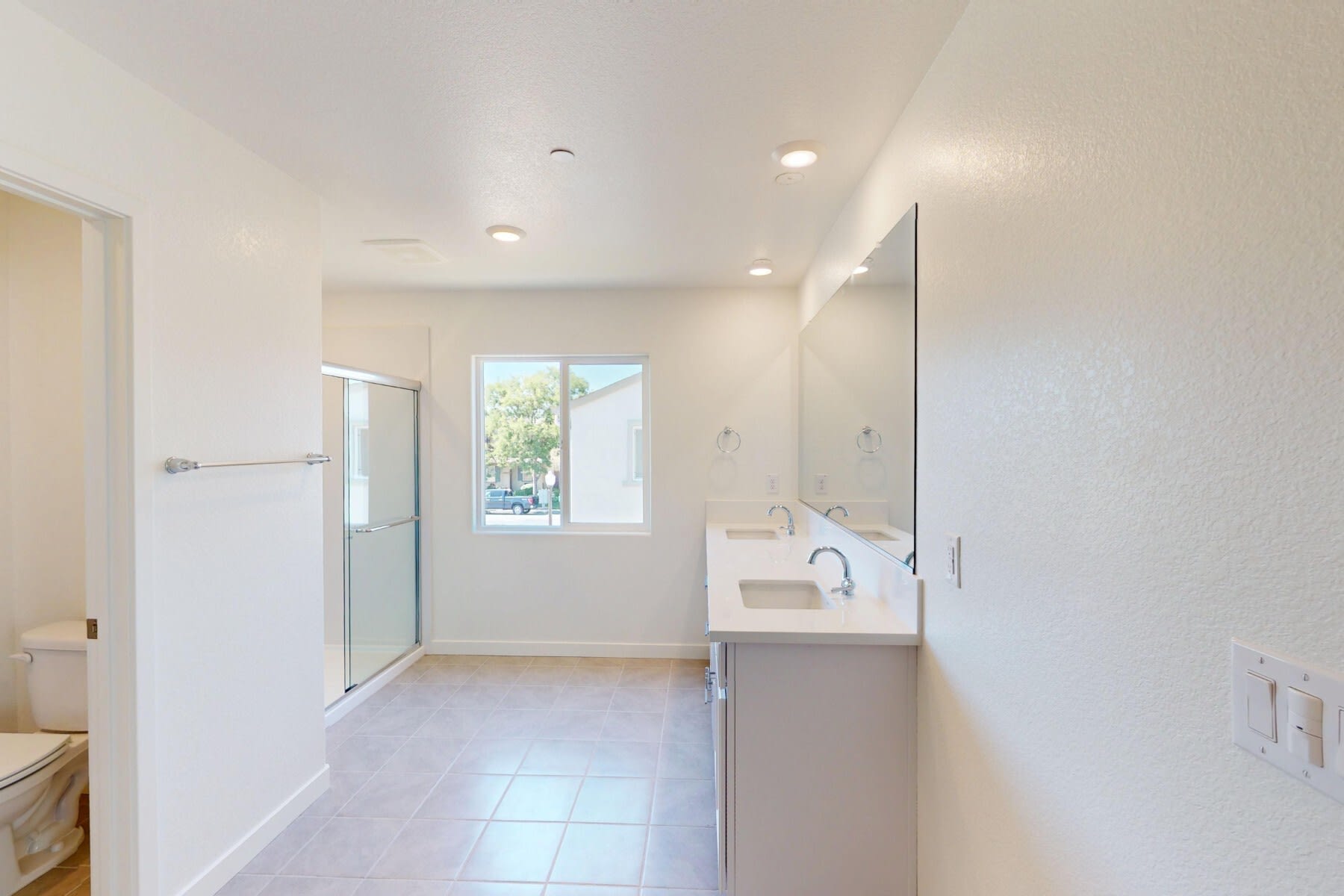 A bright and spacious bathroom with a sink, mirror, and tiled floor, leading to a hallway with a window.