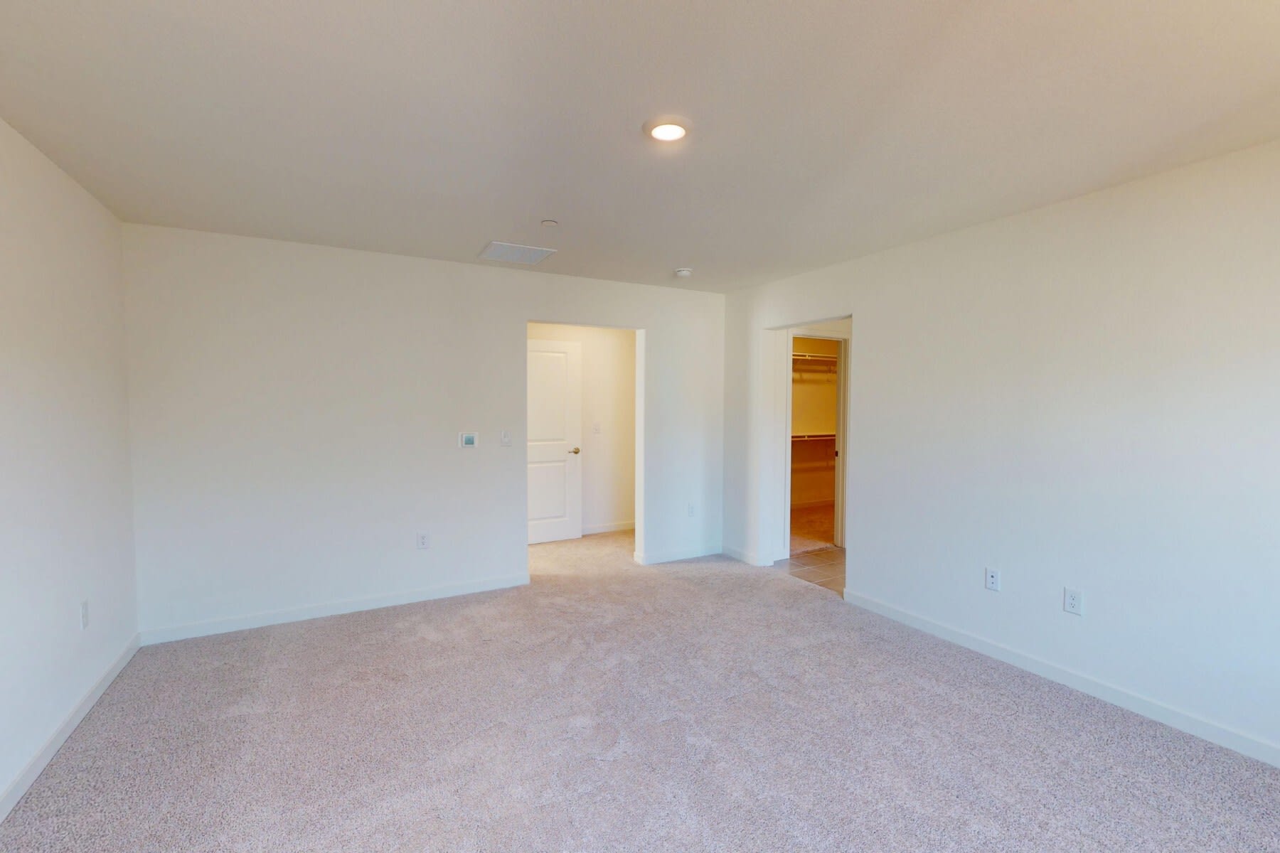 An empty, sparsely furnished room with white walls, a carpeted floor, and a doorway leading to another room.