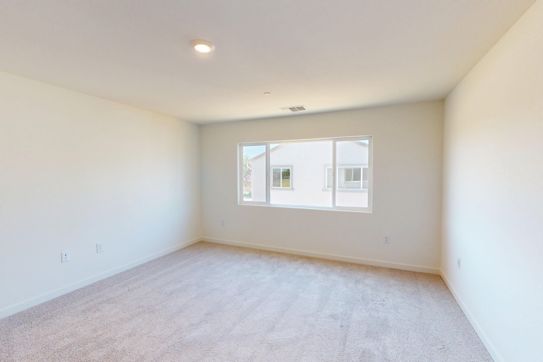 A spacious, well-lit room with a large window, a plain white wall, and a hardwood floor.