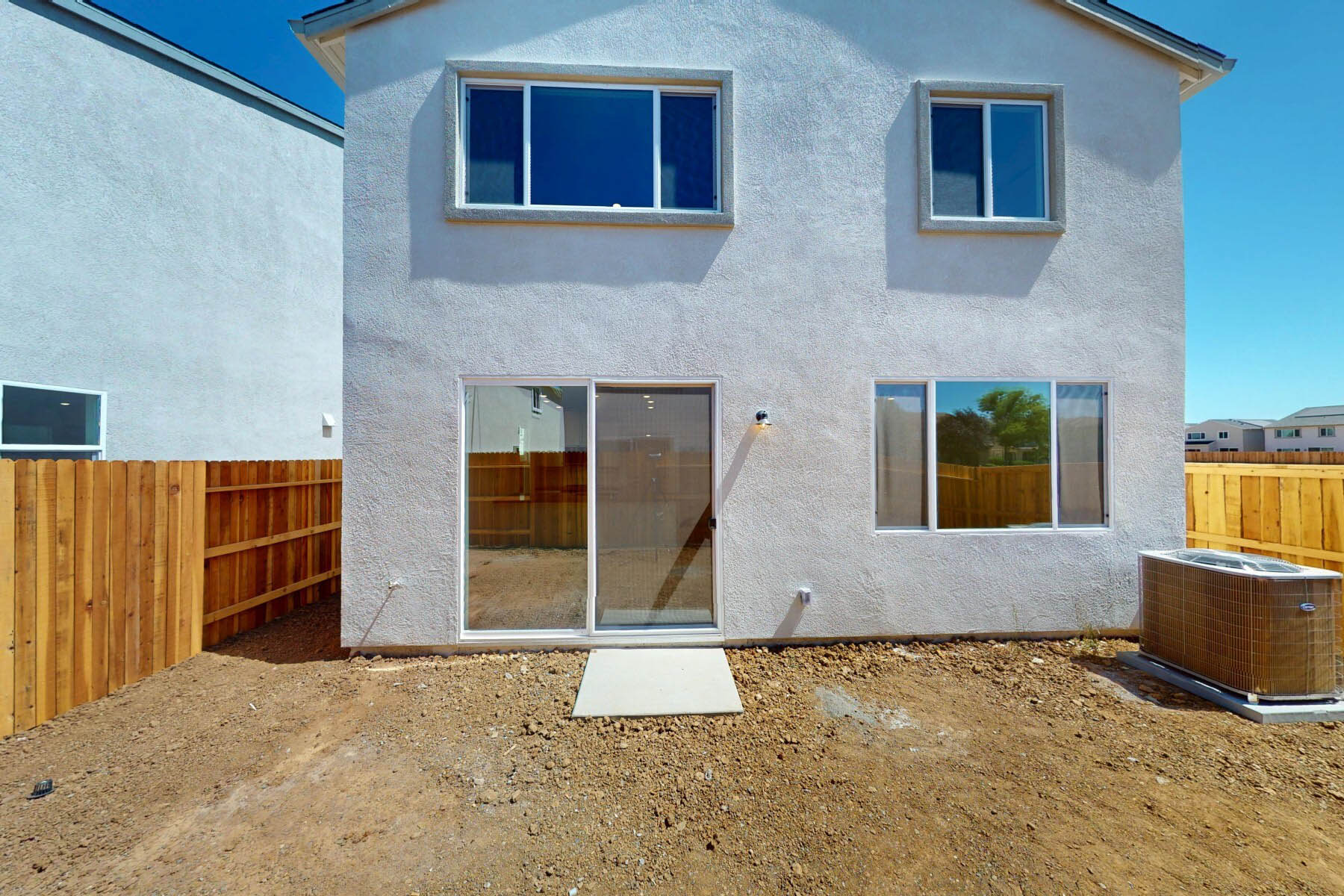A newly constructed two-story house with a wooden fence in the foreground, surrounded by a dirt yard and a clear blue sky in the background.