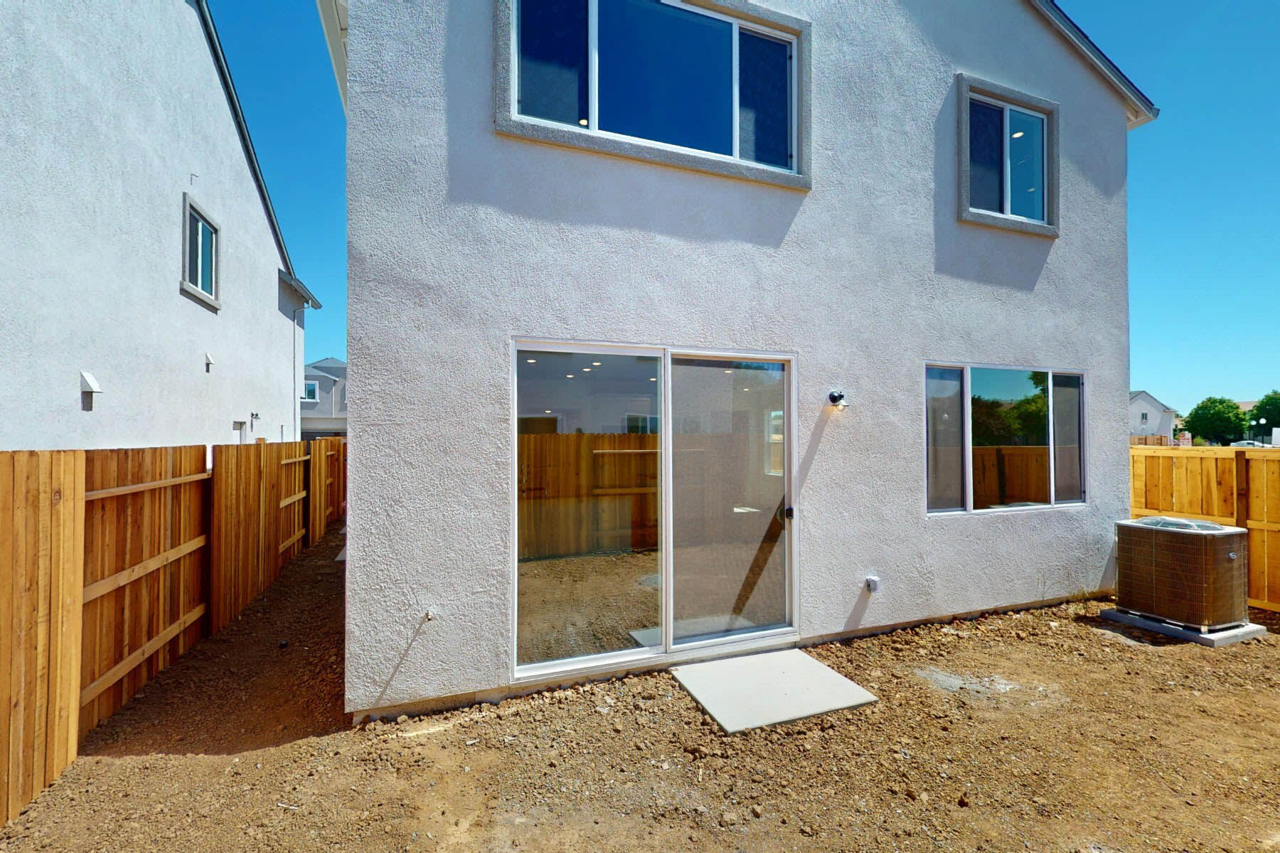 A newly constructed two-story residential building with a wooden fence in the foreground, surrounded by a dirt-covered yard and a clear blue sky in the background.