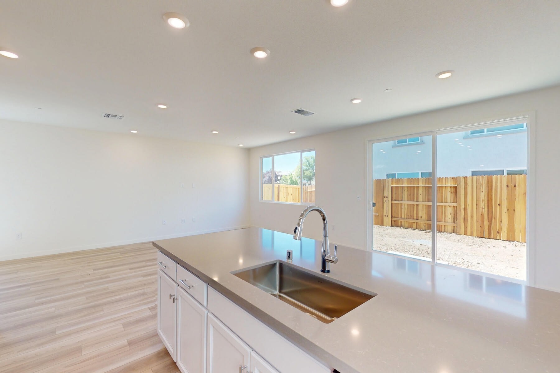 A modern, open-concept kitchen with a sleek white countertop, a stainless steel sink, and a sliding glass door leading to a fenced outdoor area.