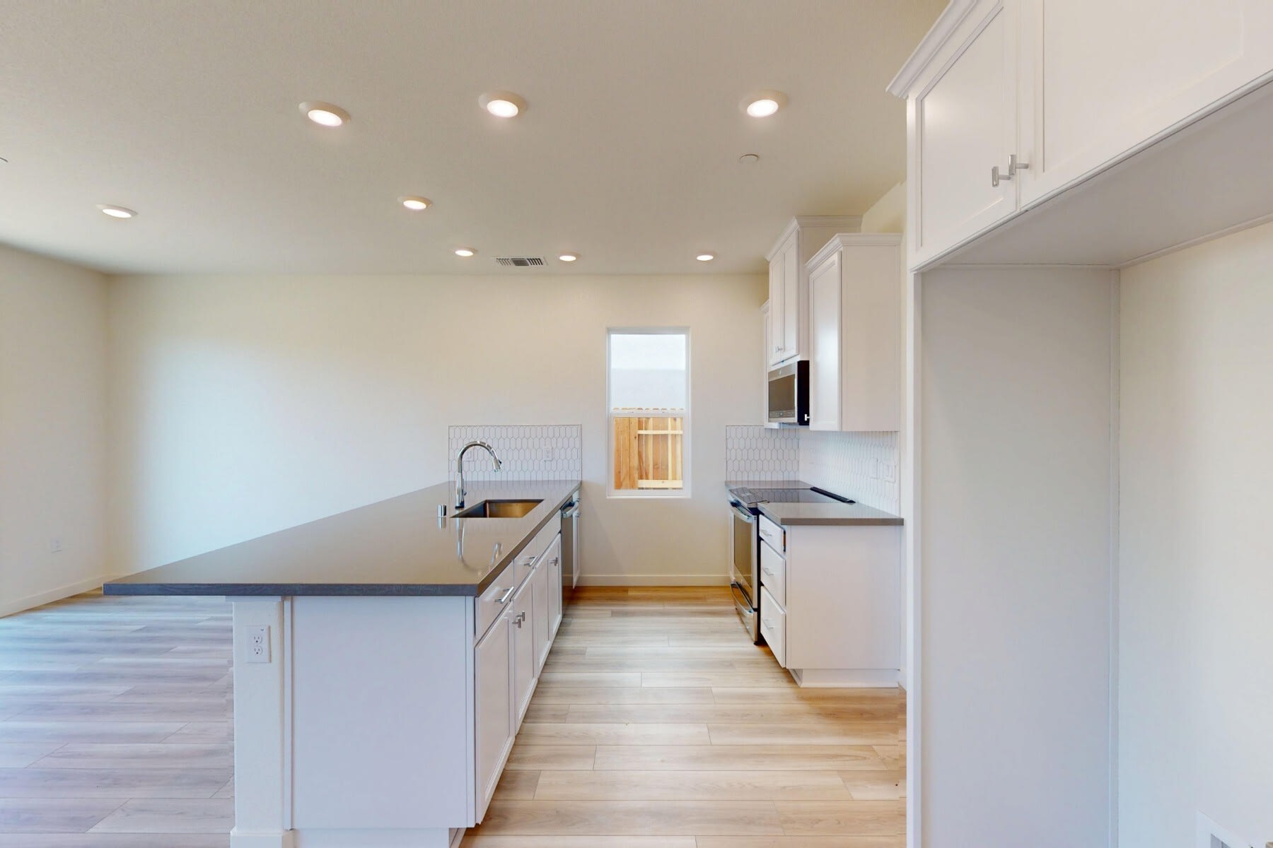 A modern, minimalist kitchen with white cabinets, a gray countertop, and hardwood floors, with a window providing natural light.