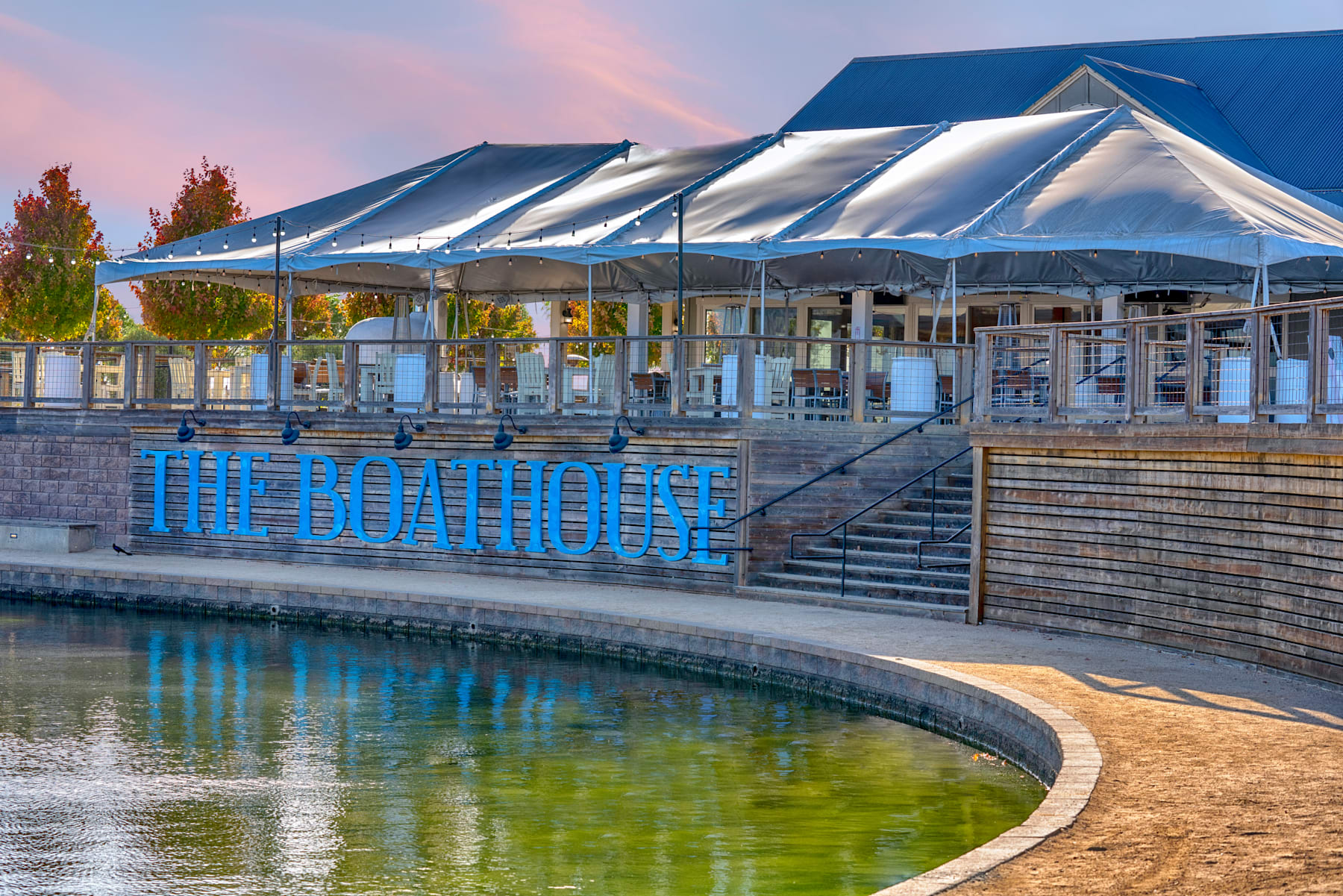 A modern boathouse structure with a curved roof and glass walls stands beside a calm body of water, surrounded by trees and a colorful sky at sunset.