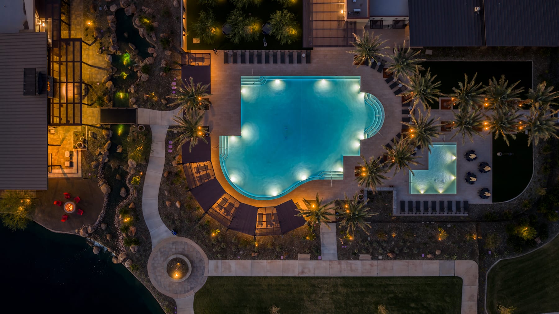 A serene, illuminated swimming pool surrounded by lush palm trees and a cozy, modern building in the background, creating a tranquil and inviting atmosphere.