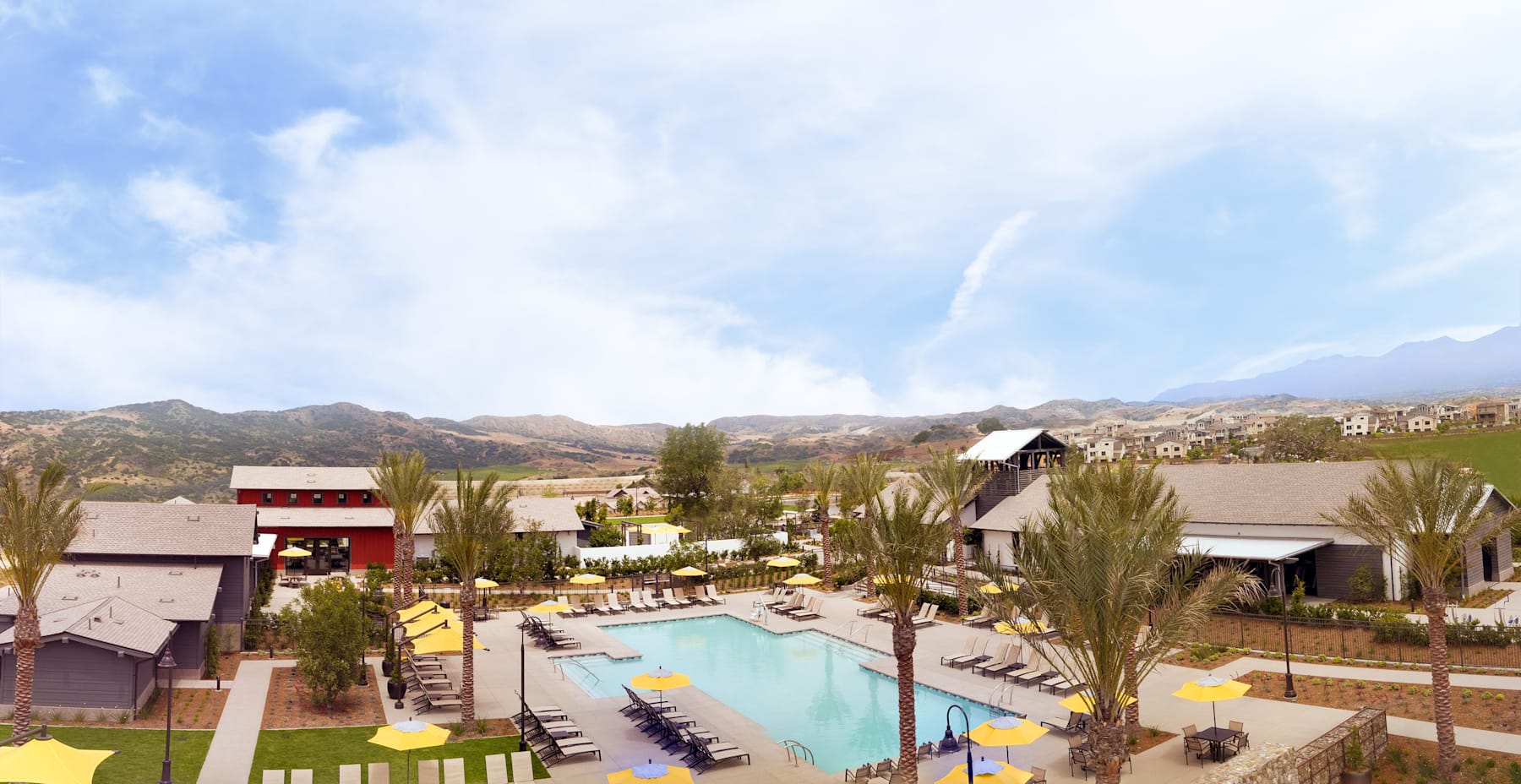 A resort-style setting with a large swimming pool surrounded by palm trees, lounge chairs, and buildings with a mountainous backdrop under a cloudy blue sky.