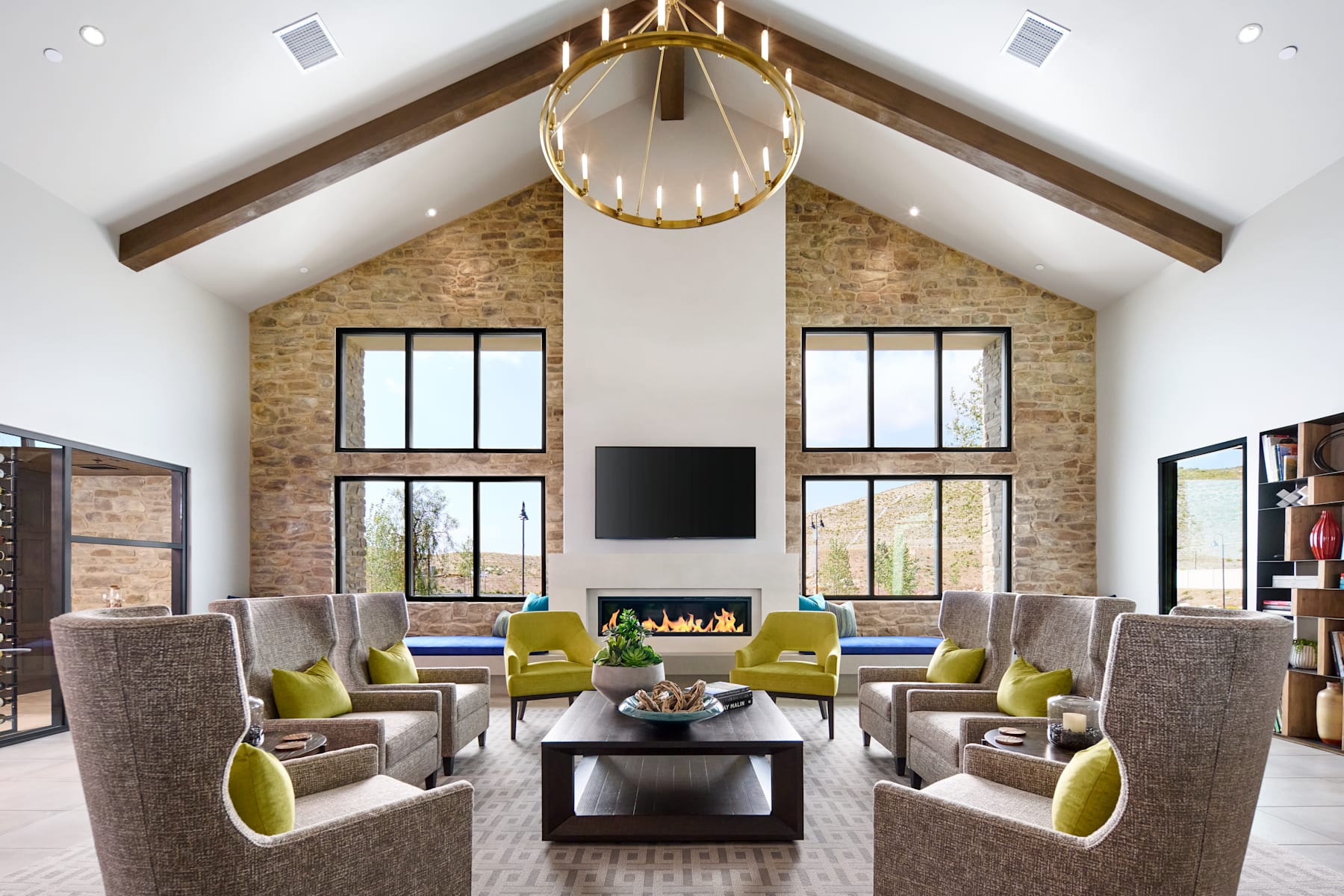 A spacious and modern living room with high ceilings, exposed brick walls, and a large fireplace surrounded by comfortable seating arrangements, including gray sofas and green accent chairs, all centered around a wooden coffee table.