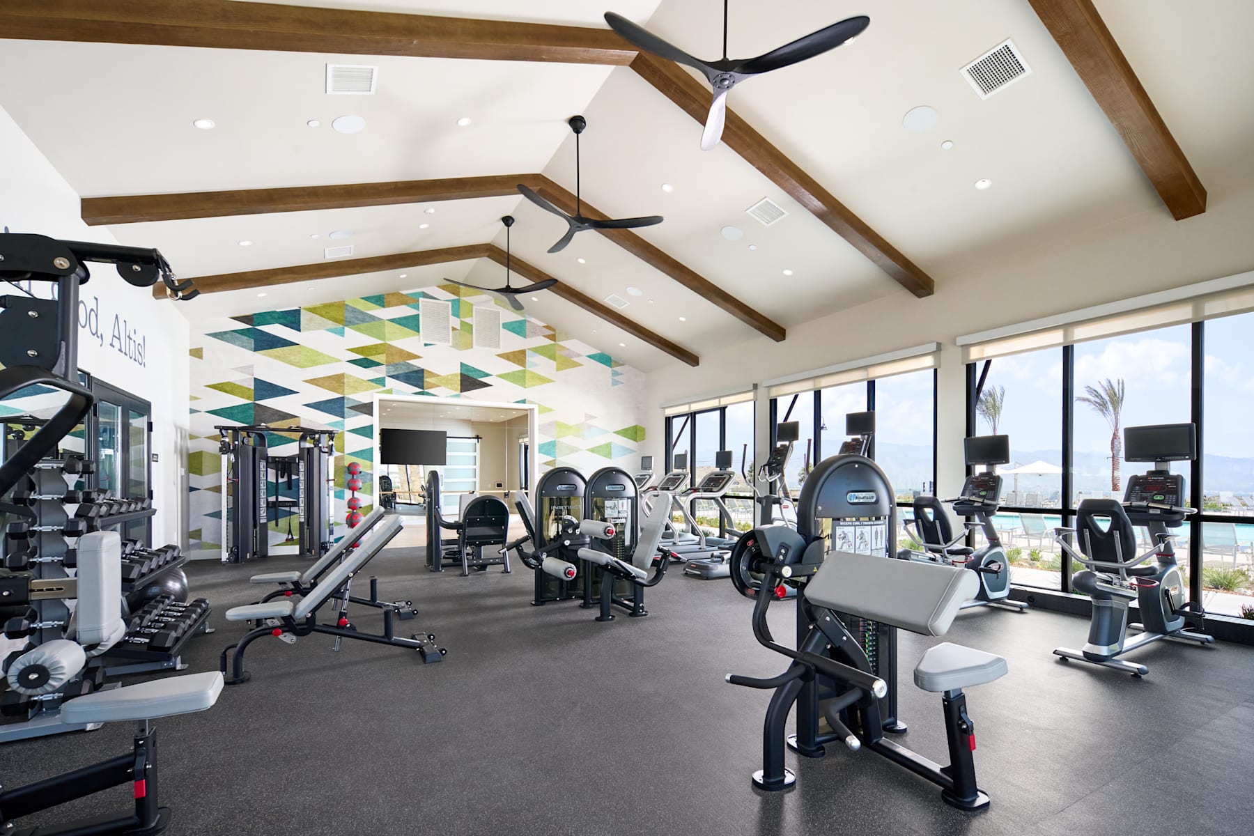 A modern and well-equipped gym with various exercise equipment, including treadmills and weight machines, set against a backdrop of wooden beams and a colorful geometric pattern on the ceiling.