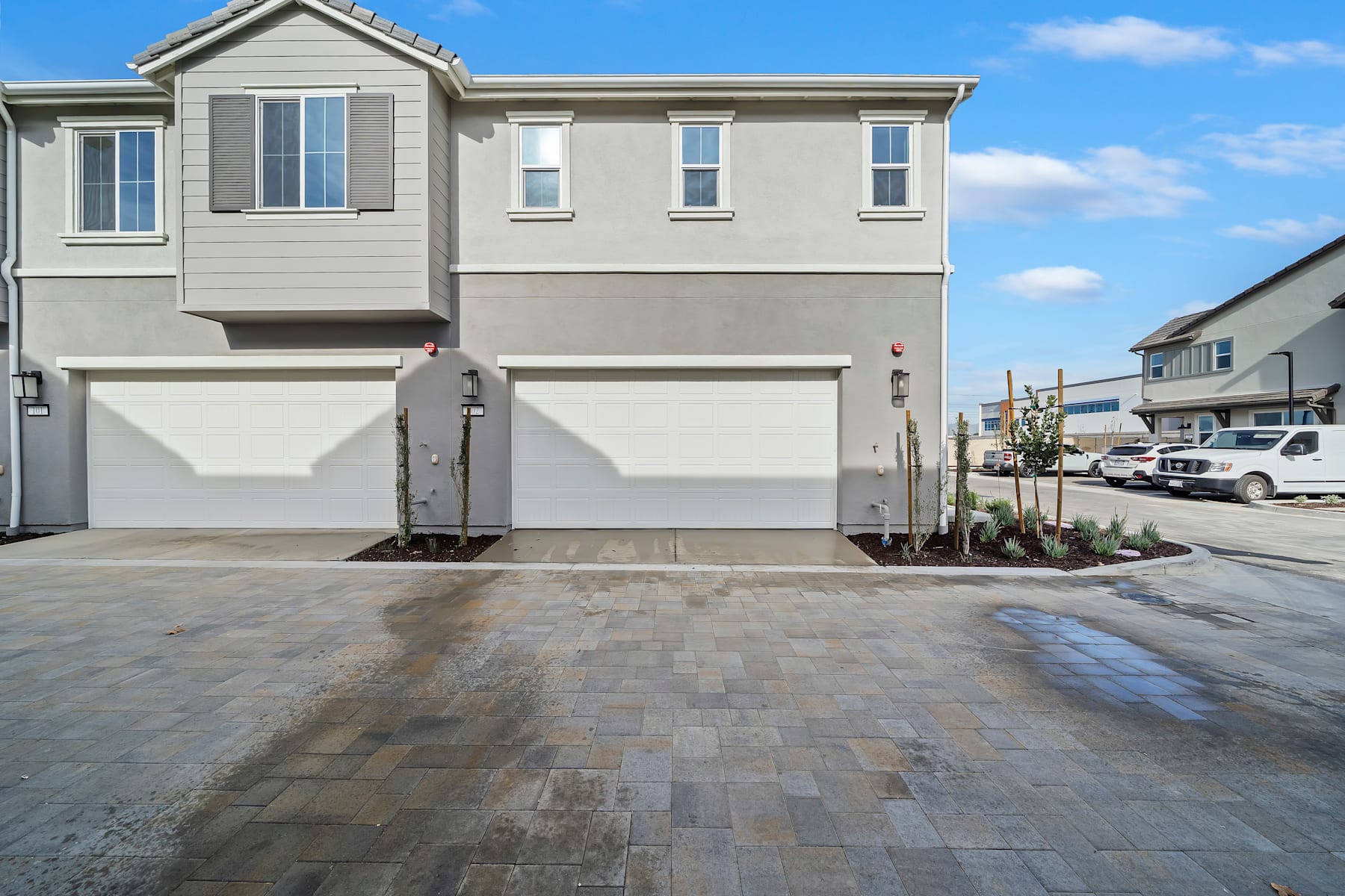 The image depicts a modern townhouse complex with a paved driveway and landscaping in the foreground, set against a clear blue sky with some clouds in the background.
