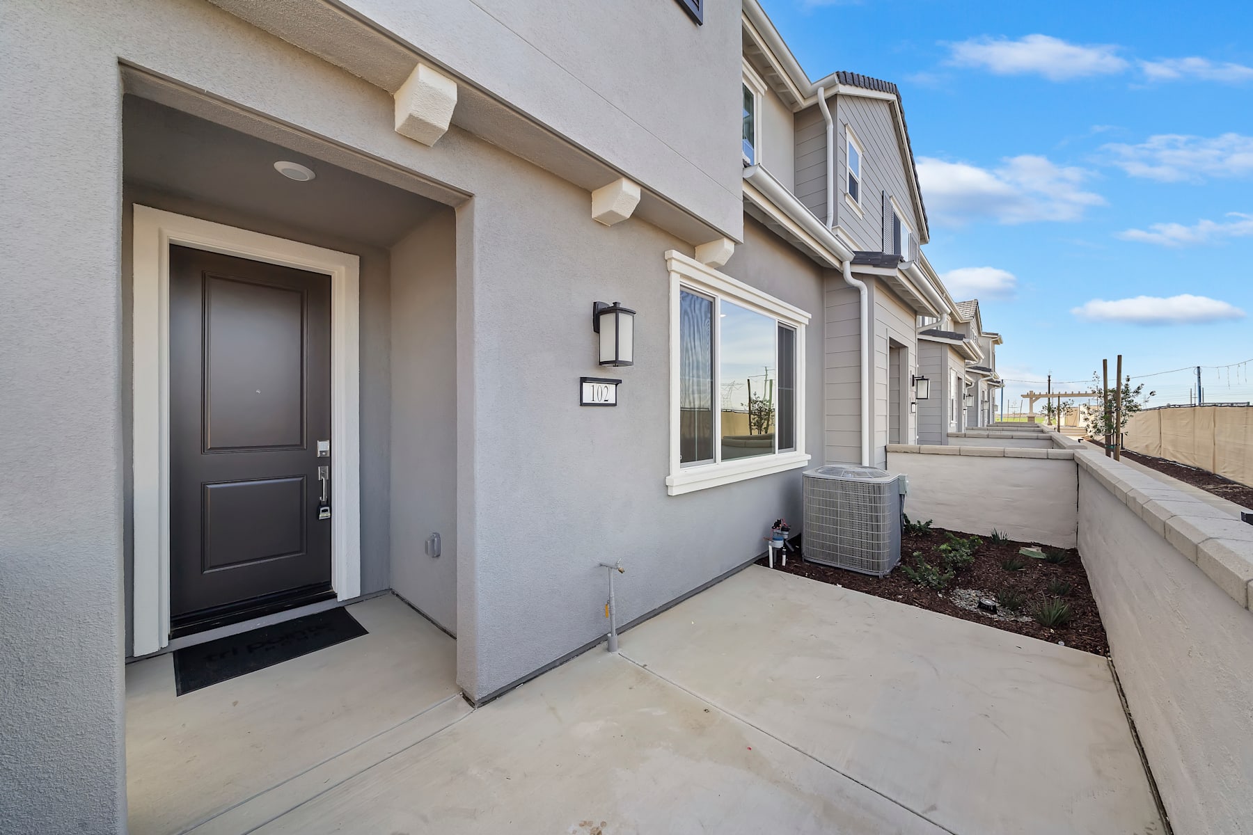 A modern, multi-story residential building with a gray exterior, a black front door, and a concrete walkway leading to the entrance, set against a clear blue sky with some clouds.